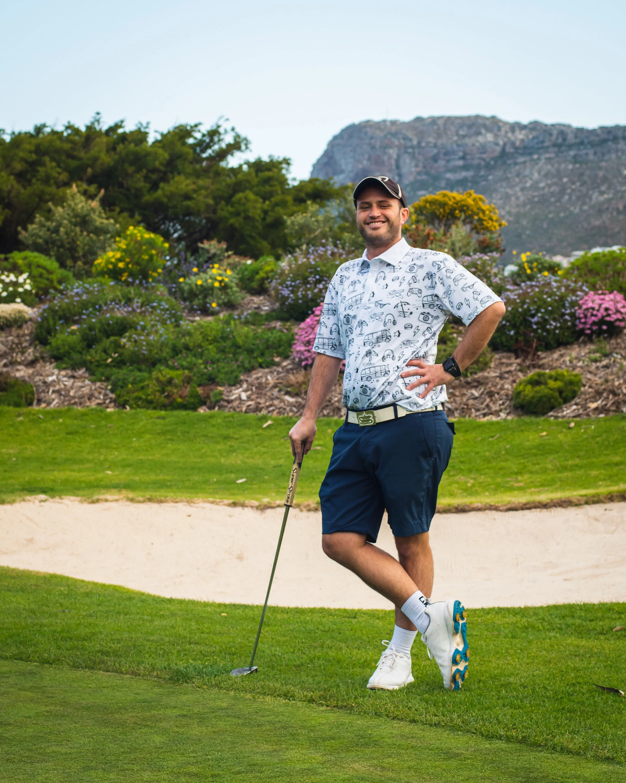 A man standing on a golf course near a sand trap, holding a golf club, smiling, wearing a white patterned shirt, blue shorts, white sneakers, and a black cap.