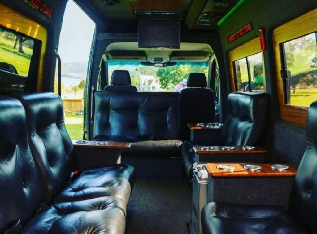 Interior of a bus or van with black leather seats, wooden armrests, and windows showing a sunny park outside.