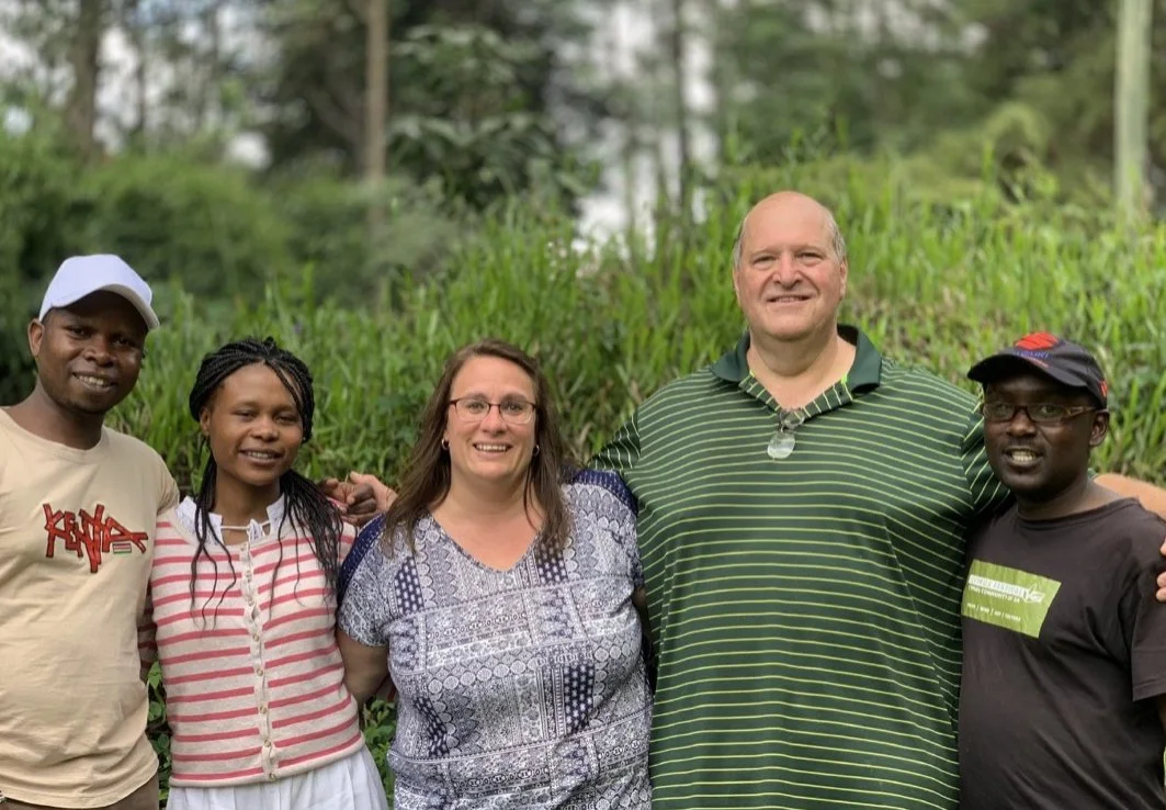 Five people standing together outdoors, smiling, in front of greenery with trees in the background.