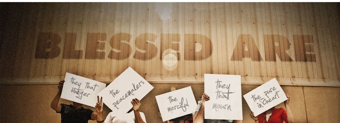 Group of people holding signs with messages such as "They that hunger," "The peacemakers," "The merciful," "They that mourn," and "The pure in heart" in front of a wooden wall with the words "Blessed Are" painted on it.
