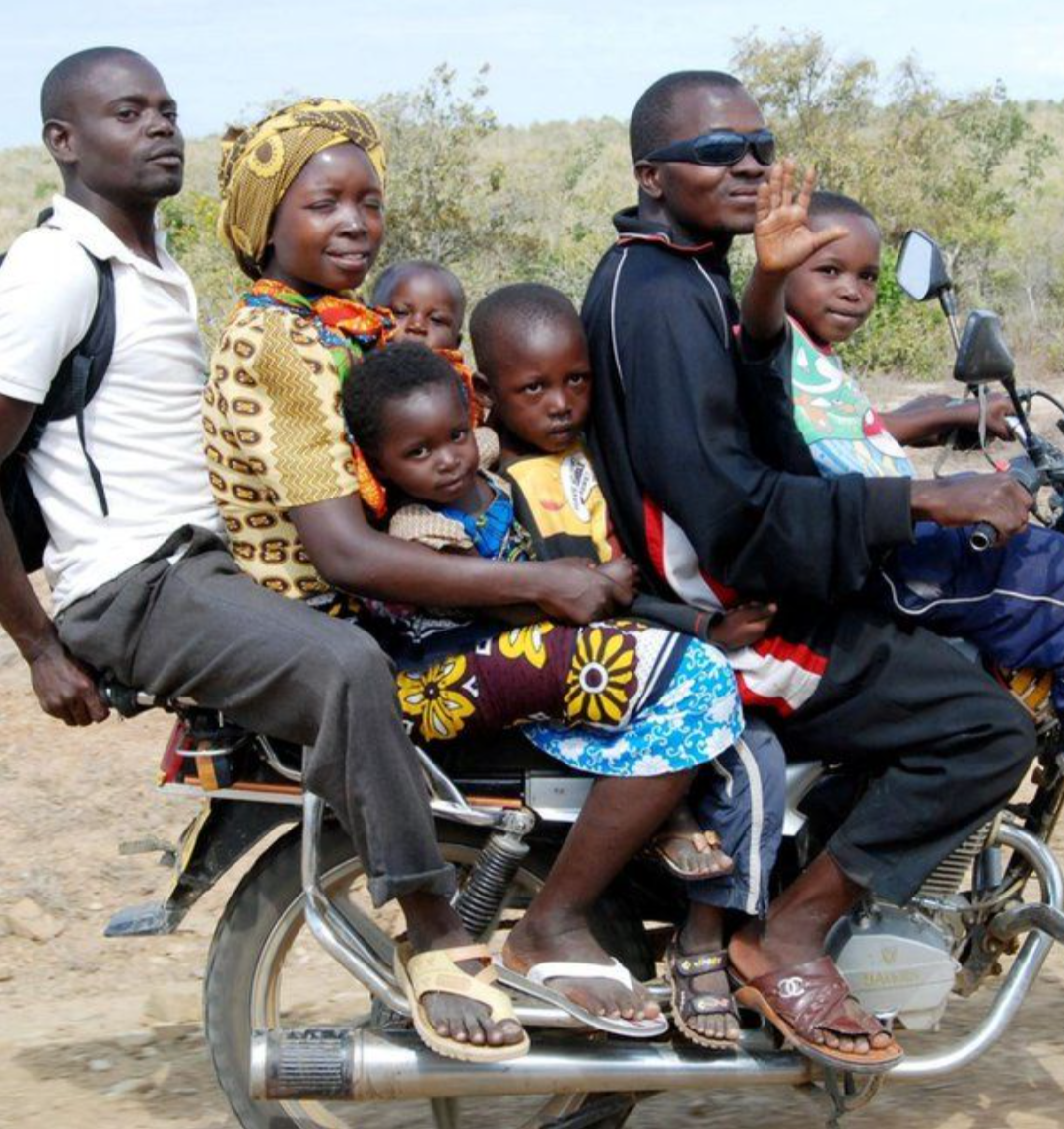 A group of people riding on a motorcycle in a rural area, including children and adults, some of whom are waving or looking at the camera.