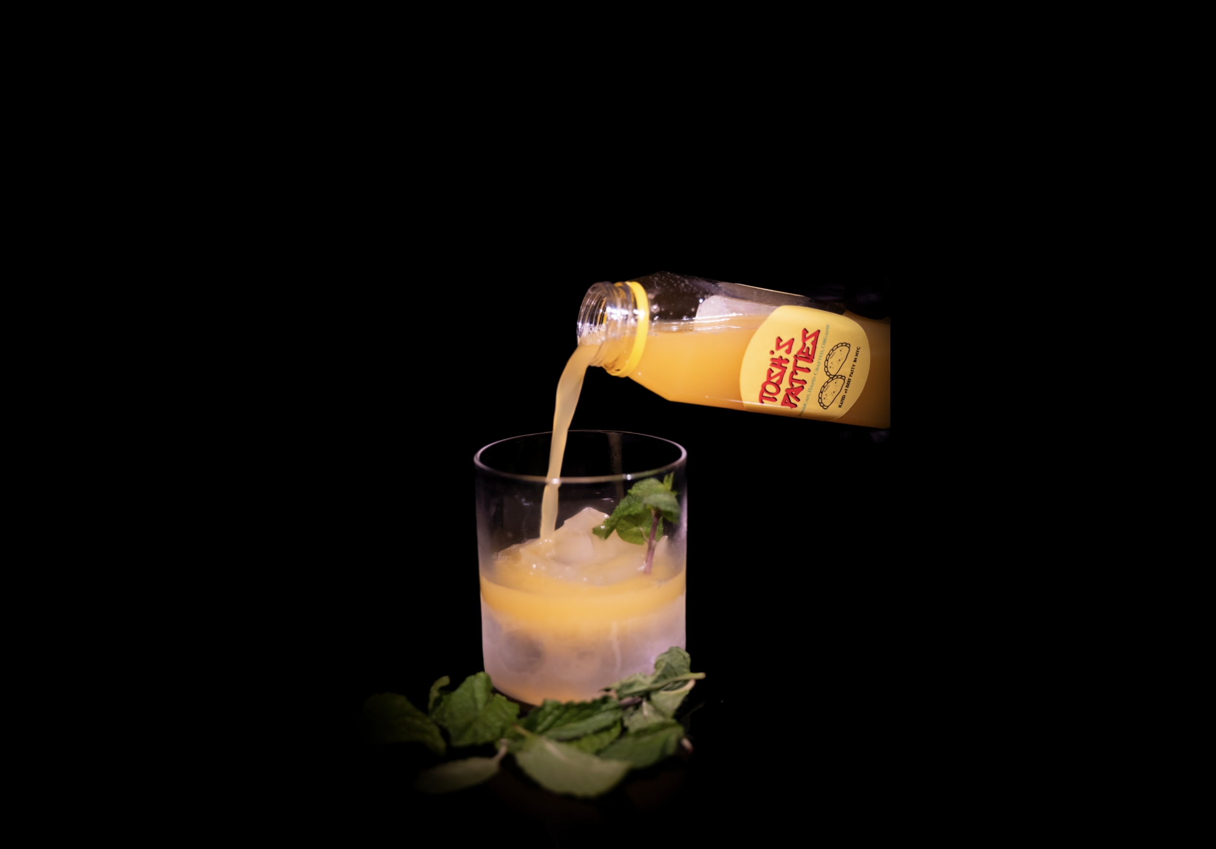 Toys R' Us-branded orange juice being poured into a glass with ice and mint leaves, with some mint leaves on the surface, against a black background.