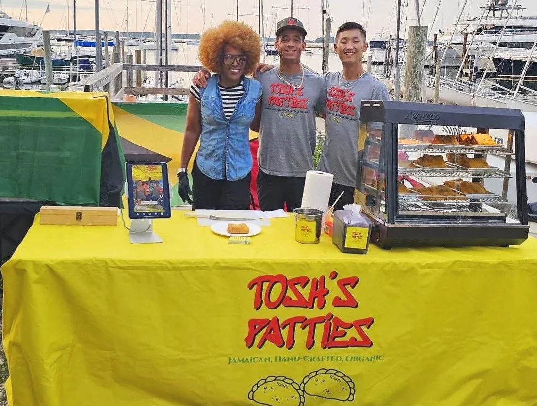 Three people standing behind a yellow table with a sign that says 'Tosh's Fatties' at a marina. The table has a display case of food, a plate with food, and a digital cash register. The marina background includes boats and docks.