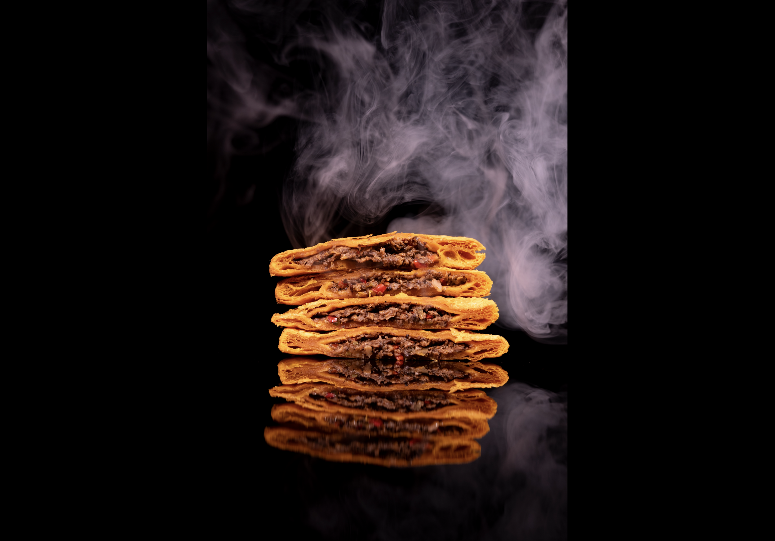 Stack of four spicy beef empanadas with visible red chili peppers, emitting smoke against a black background, with reflection below.