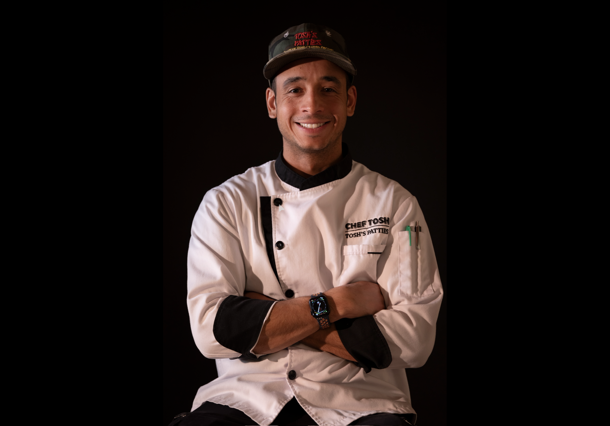 A smiling male chef in a white chef's coat with black accents, wearing a black cap, with arms crossed, standing against a dark background.