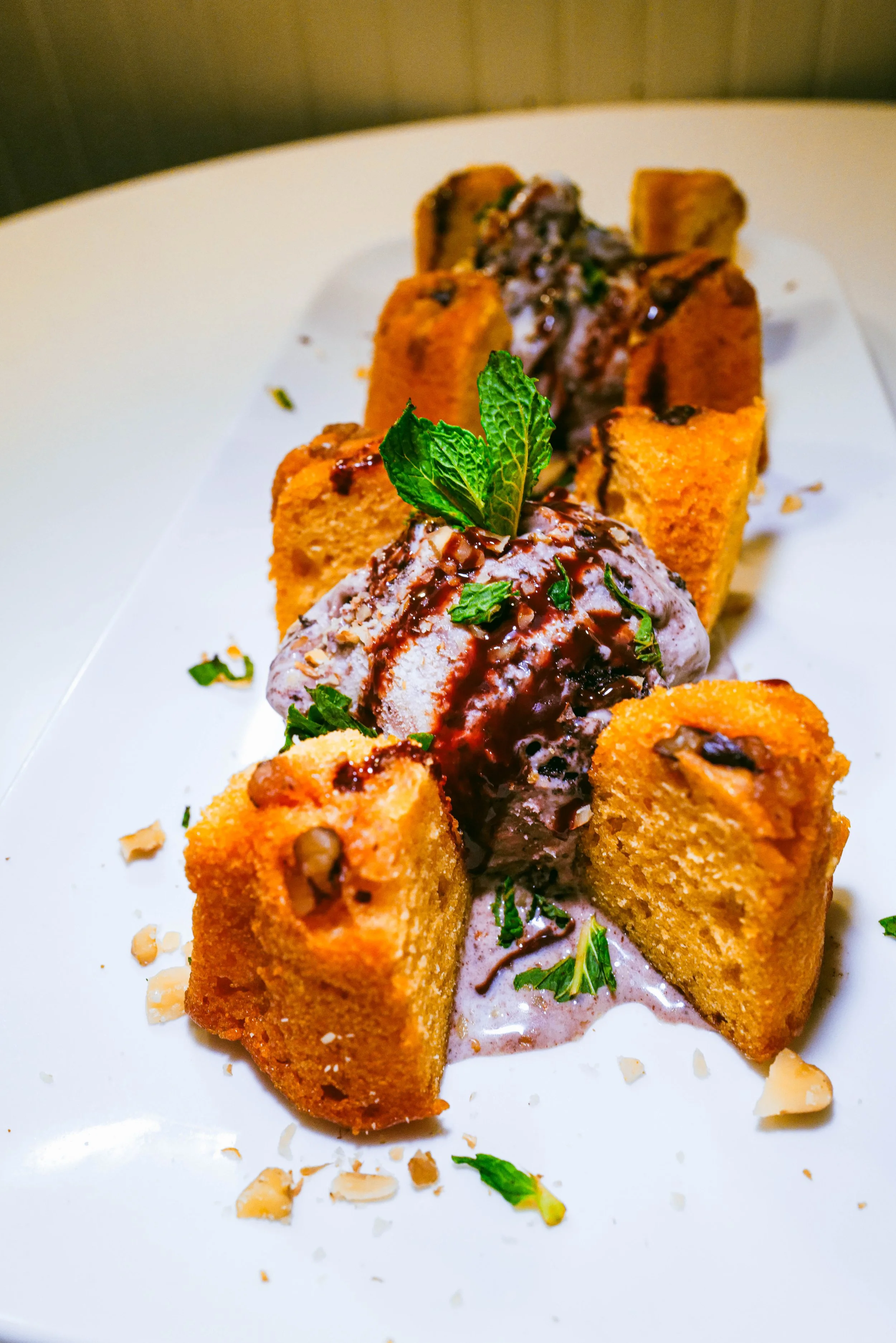Fried dessert squares topped with ice cream, chocolate sauce, chopped nuts, and fresh mint leaves on a white plate.