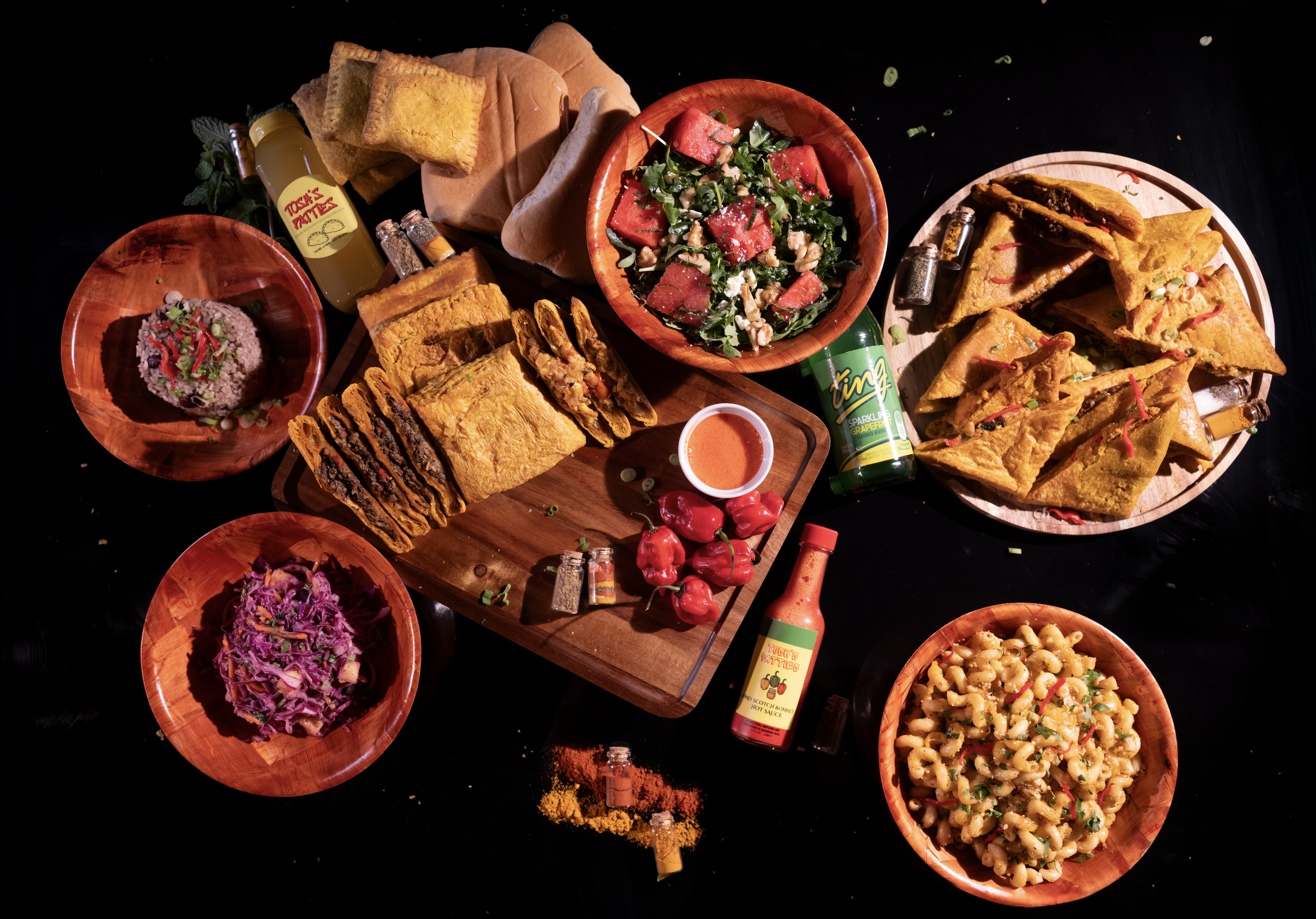 A variety of Mexican dishes and condiments on a black background, including tacos, tortilla chips, salsa, salad, macaroni salad, and rice, with bottles of hot sauce and seasoning.