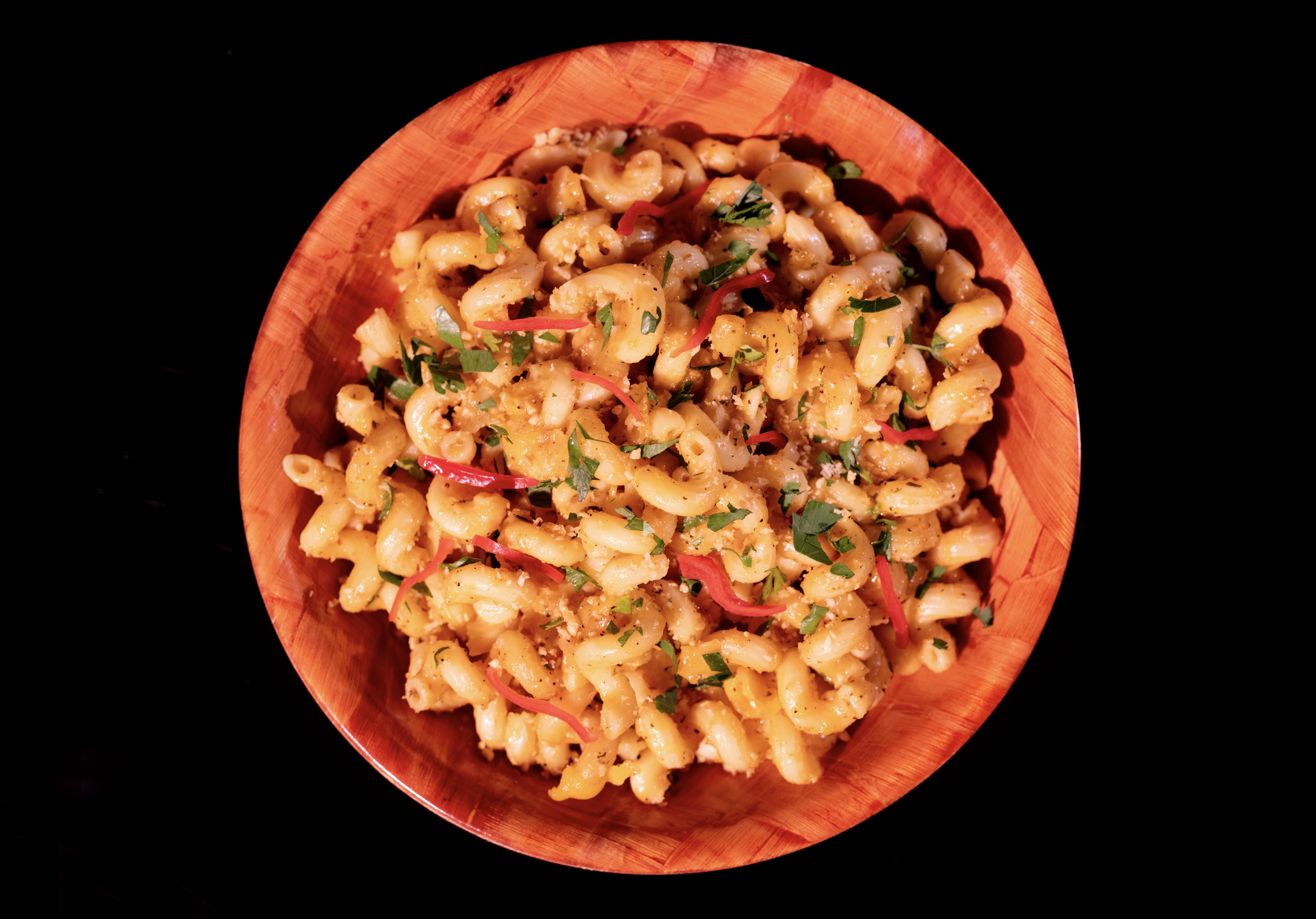 A wooden bowl filled with cooked pasta salad garnished with chopped herbs and thin slices of red chili pepper, placed on a black surface.