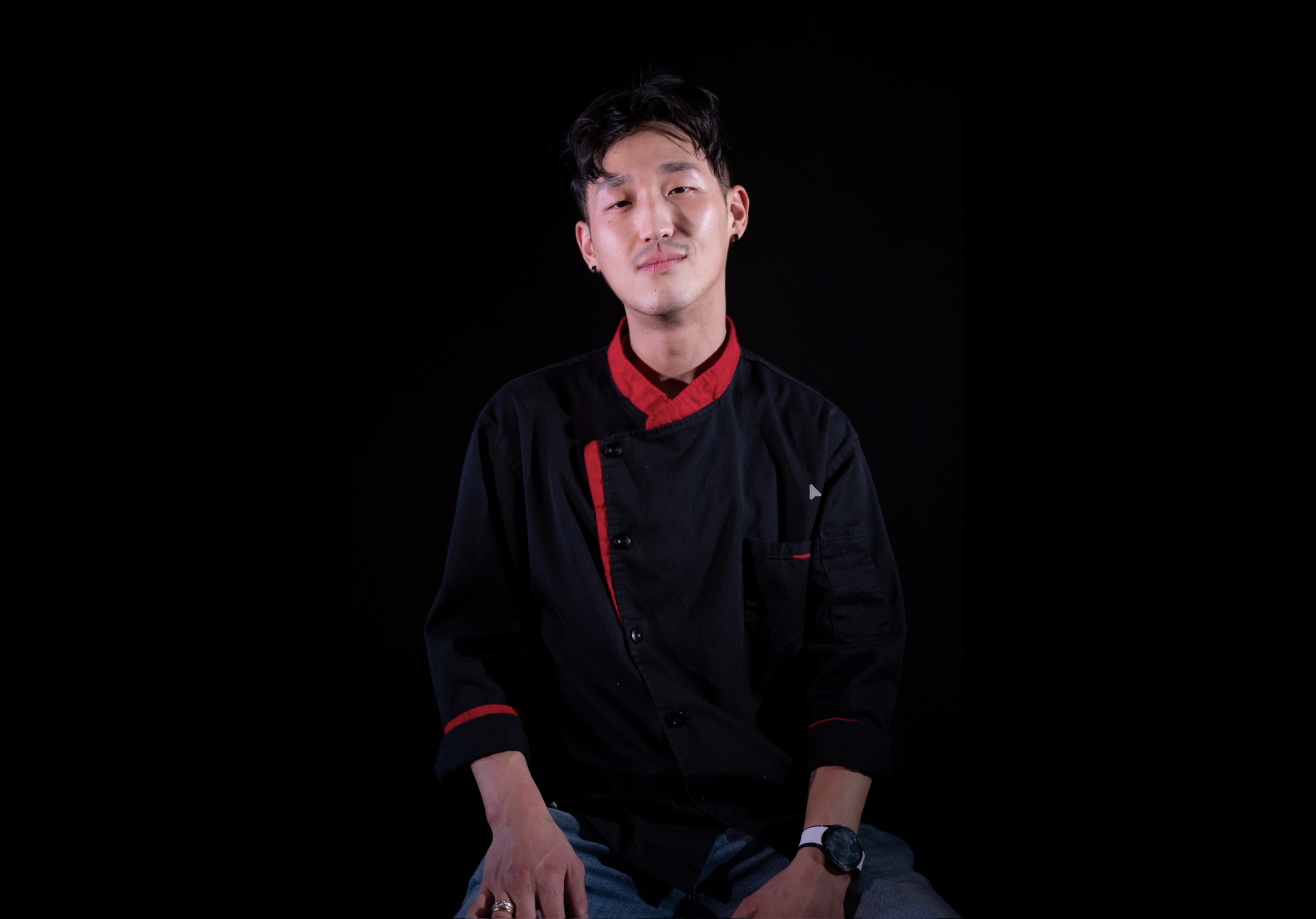 A young man in a black chef's jacket with red accents, sitting against a black background.
