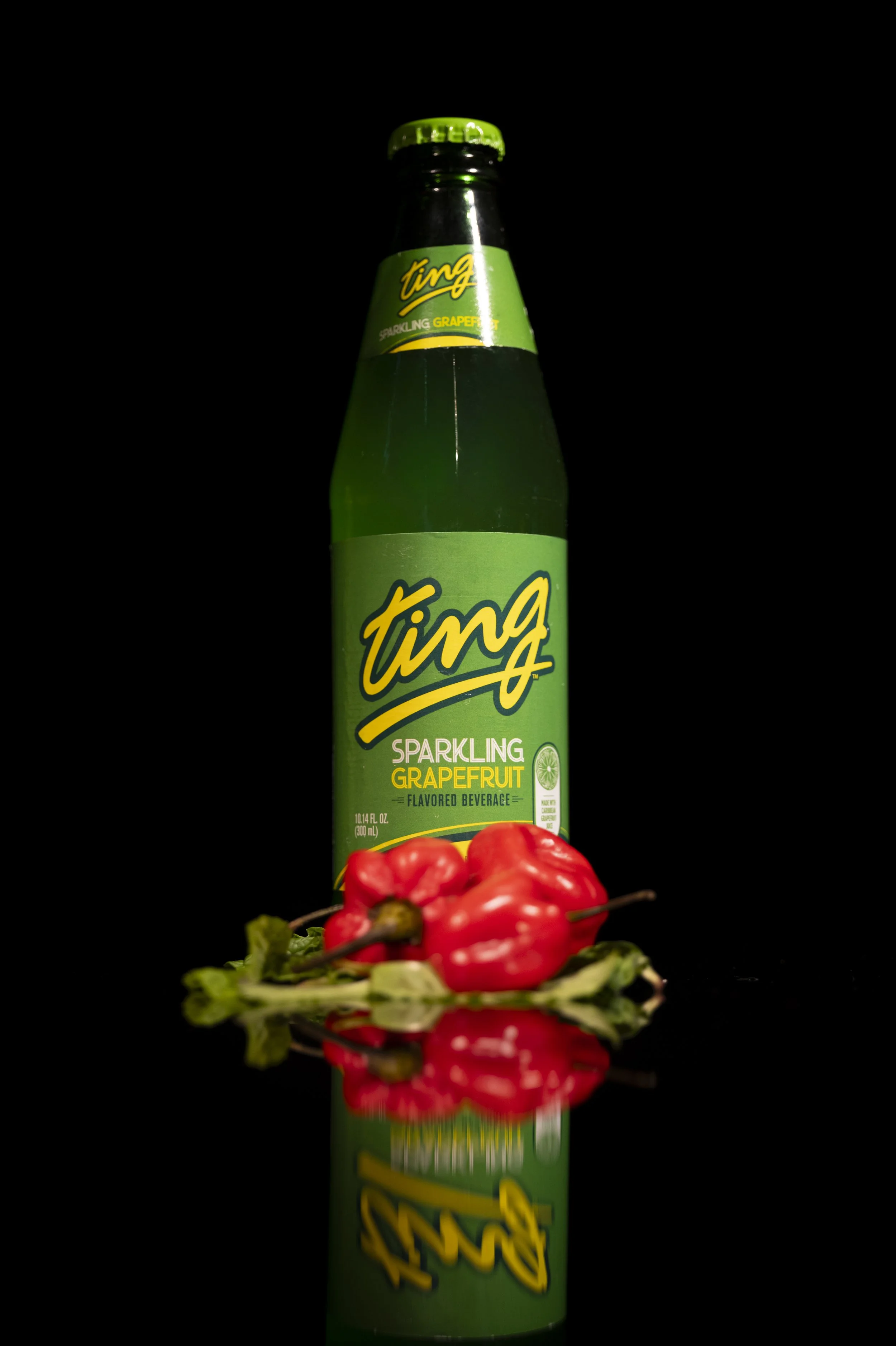 A green glass bottle of Ting sparkling grapefruit beverage with a yellow and green label, placed behind two red habanero peppers and some green leaves, against a black background with their reflection below.