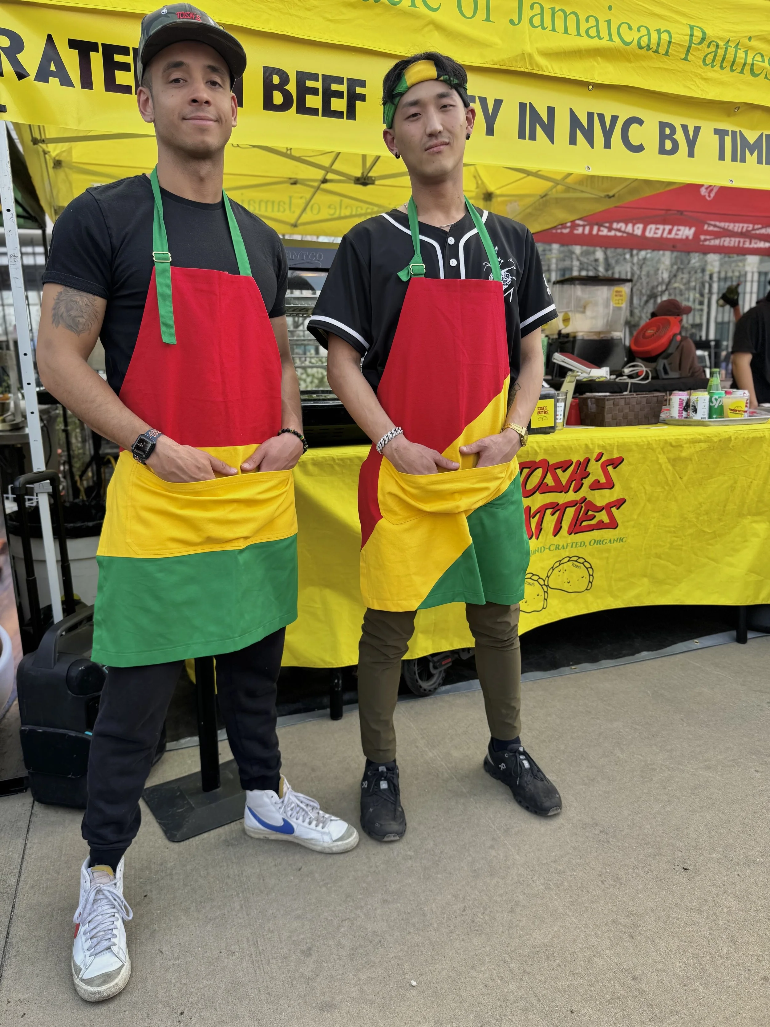 Two men standing at a booth, wearing aprons with red, yellow, and green colors. The booth has a yellow banner with green and black text. One man is wearing a black t-shirt, black pants, white sneakers, and a black cap. The other man is wearing a blac