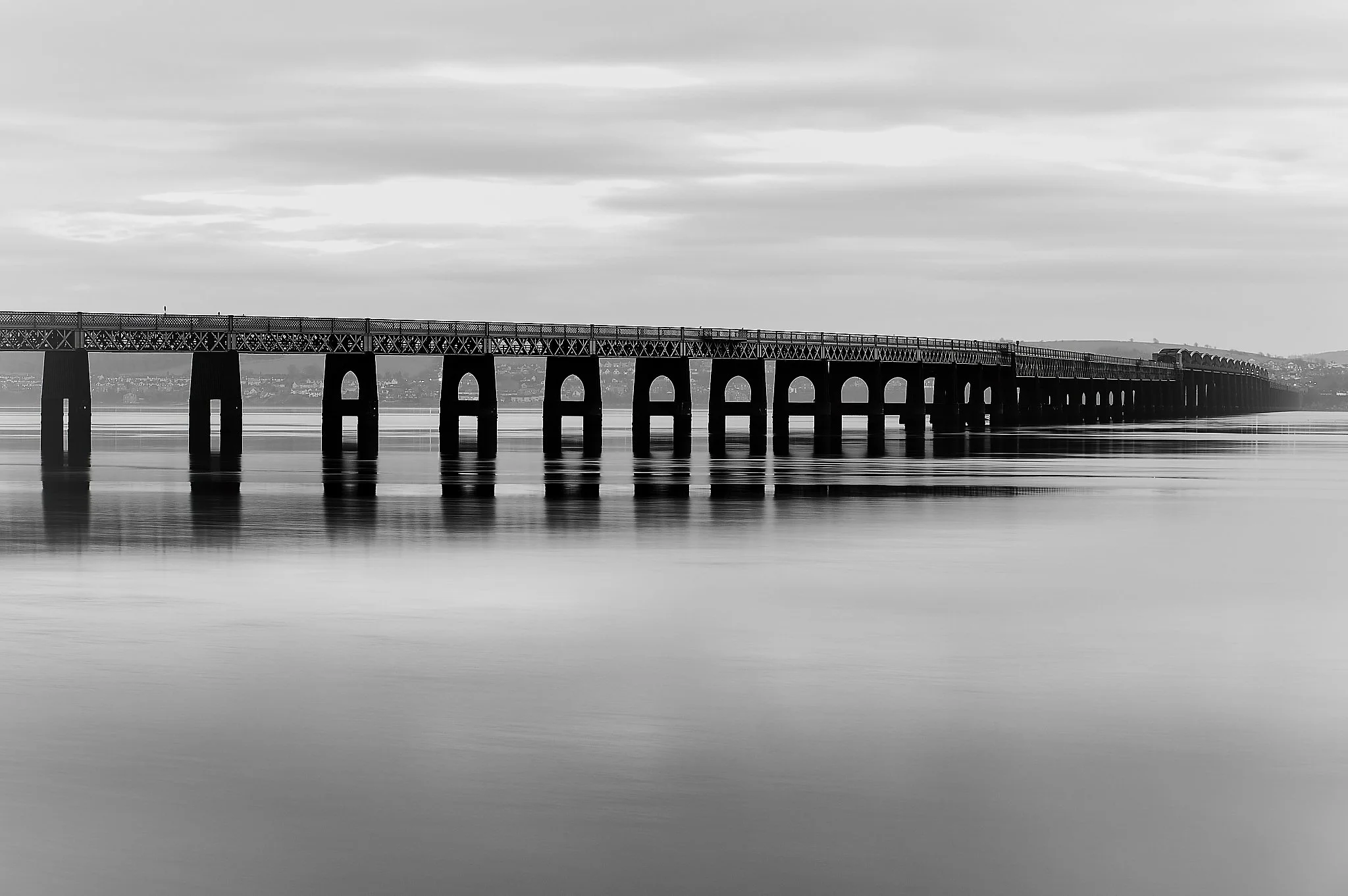 The Tay Rail Bridge stretches across the water in a perfect rhythm of arches and pillars, its geometry sharpened by the clarity of black and white. The calm surface of the river mirrors the structure above, softening the steel with a quiet sheen and 