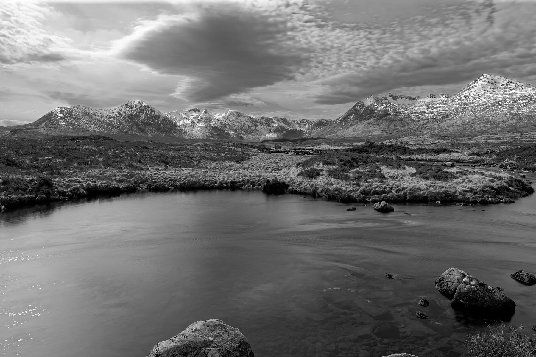 A moment of absolute quiet settles over Rannoch Moor, where water, rock, and sky meet in a rare harmony. This beautifully bleak landscape reveals its raw Highland character — the glass‑smooth lochan reflecting a world carved by ice, while distant sno