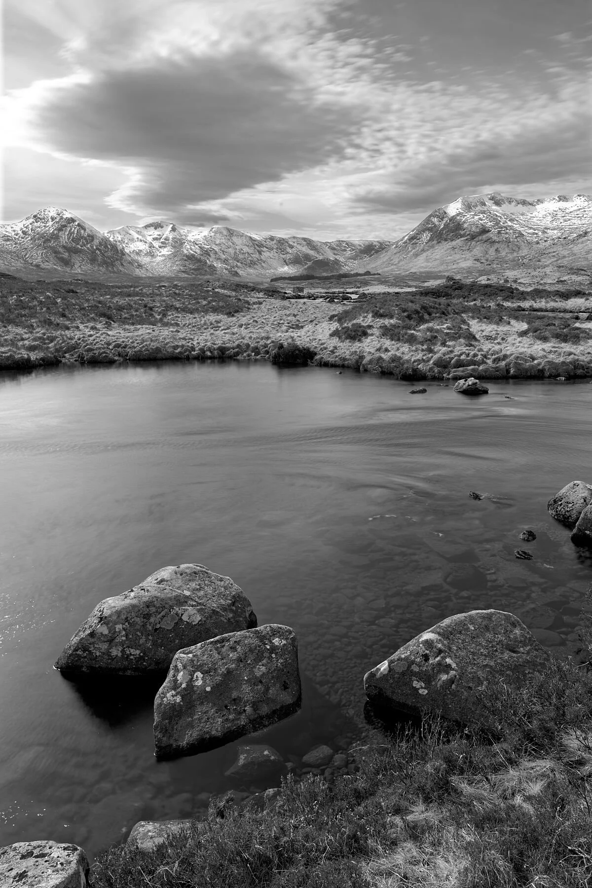 A moment of absolute quiet settles over Rannoch Moor, where water, rock, and sky meet in a rare harmony. The beautifully bleak landscape reveals its raw Highland character — the glass‑smooth lochan reflecting a world carved by ice, while distant snow