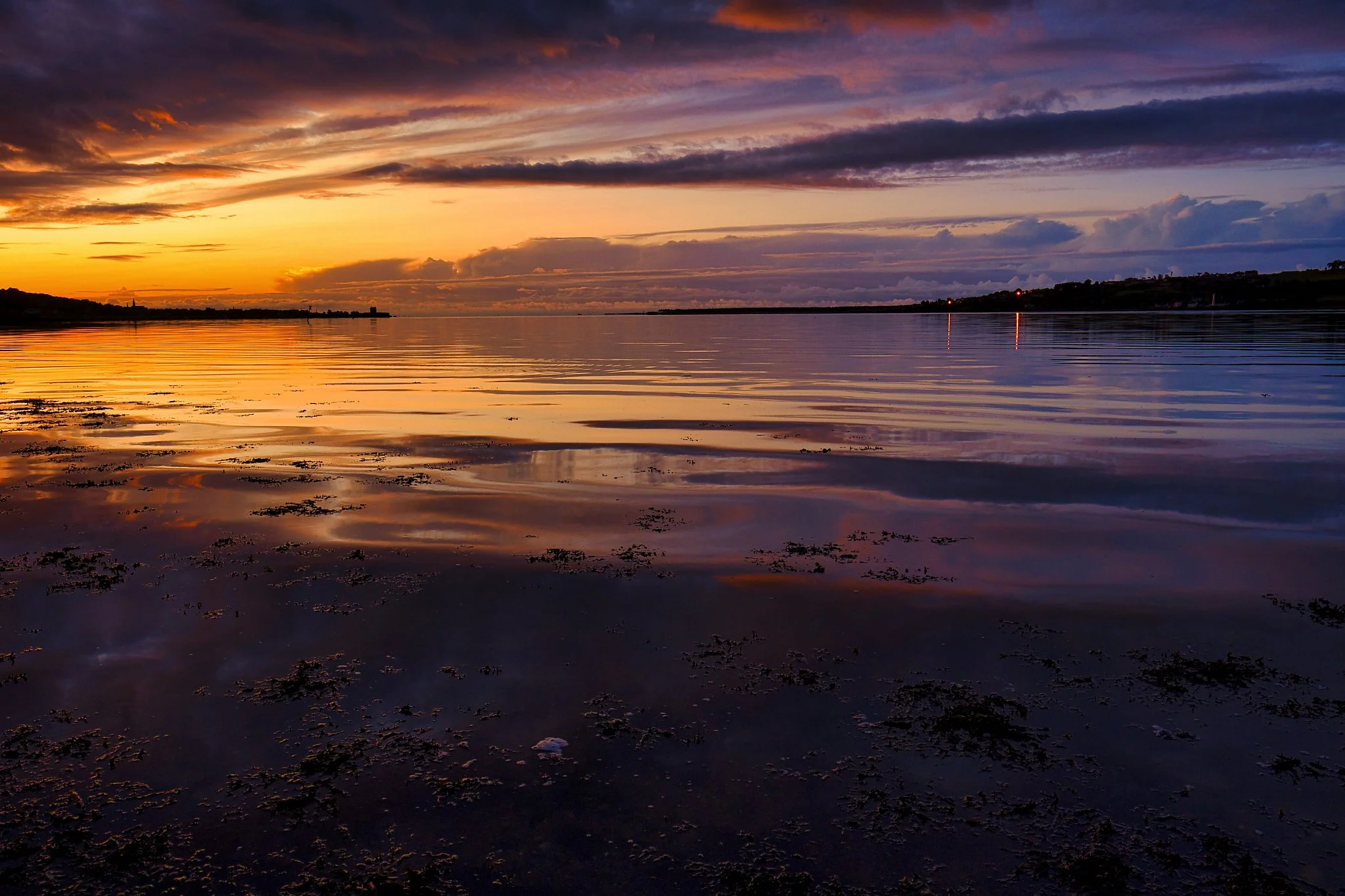 The first light of morning spreads across the Tay, painting the sky in deep oranges, soft purples, and fading blues. The water lies calm and reflective, mirroring the colours above with a quiet clarity that makes the whole scene feel suspended in tim