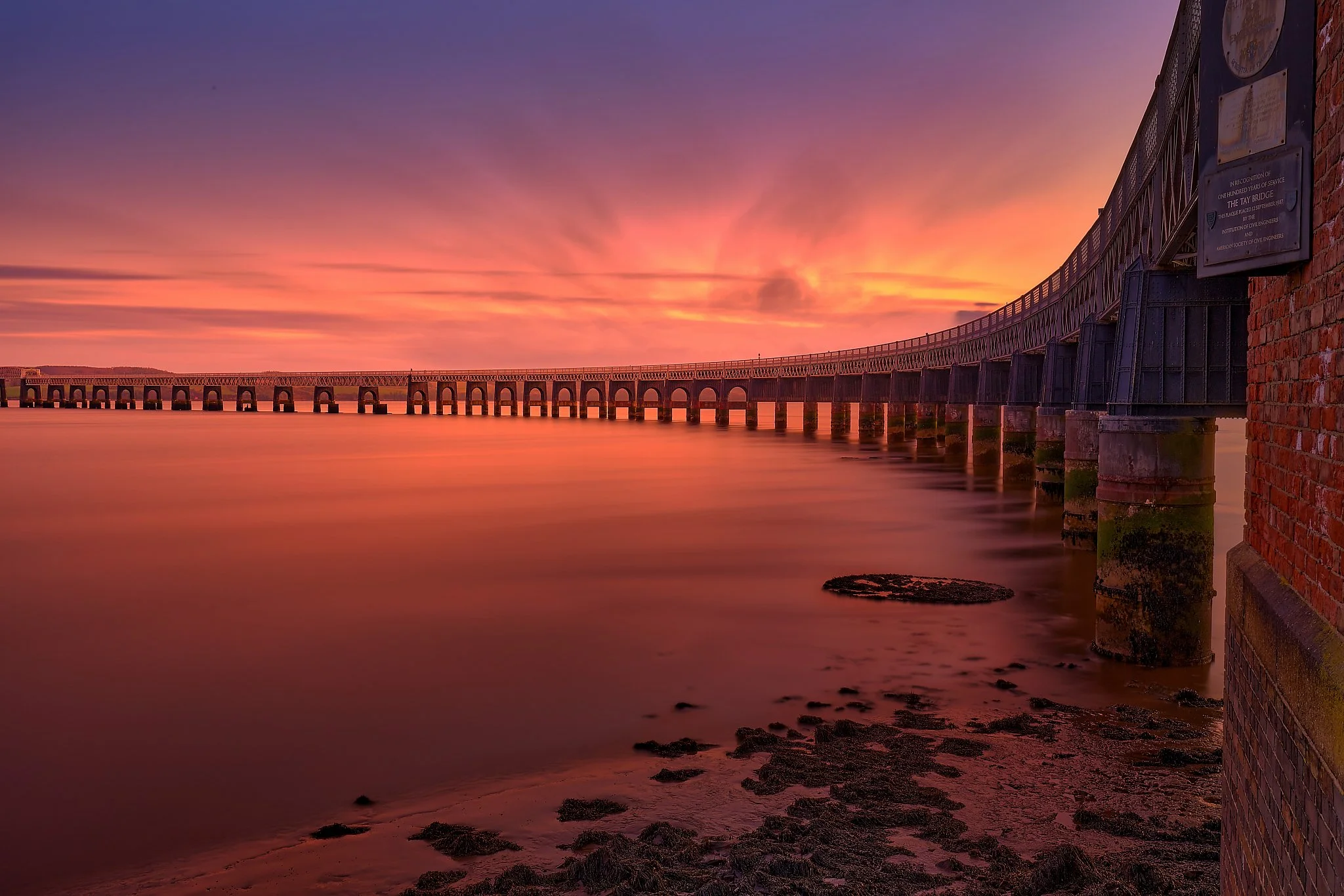 The Tay Rail Bridge stretches across the river like a quiet rhythm, each pillar catching the last warmth of the day as the sun sinks behind the horizon. Evening light washes the sky in deep oranges, soft pinks, and fading purples, mirrored in the sti