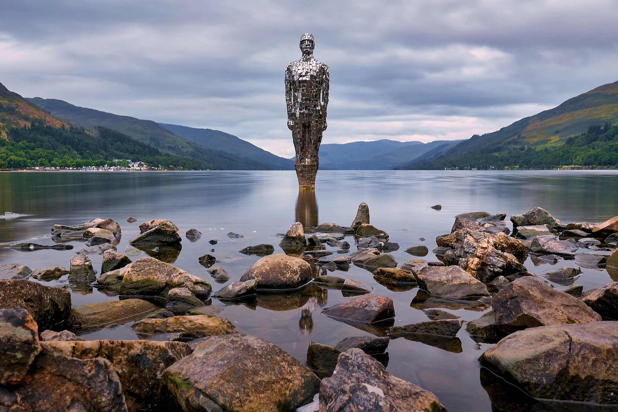 A solitary figure rises from the calm surface of Loch Earn, its mirrored panels catching the soft Highland light and reflecting the world around it. The sculpture stands motionless in the water, a quiet presence framed by rocky shores, rolling green 