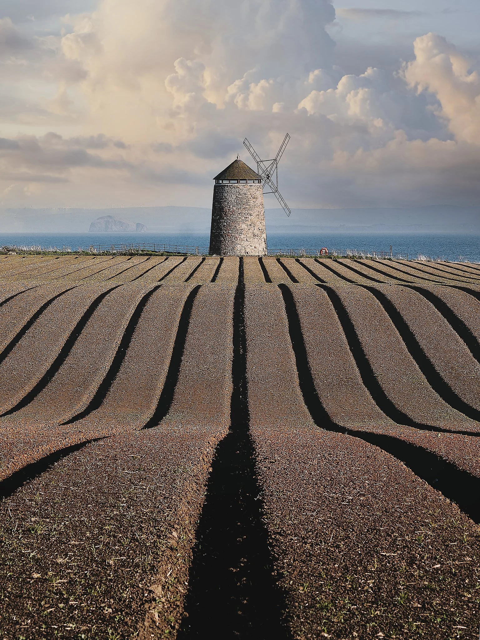 The old windmill of St Monans rises from the landscape like a quiet sentinel, its stone walls weathered by centuries of coastal wind. In the foreground, the freshly ploughed field forms a series of bold, sweeping lines that draw the eye directly towa