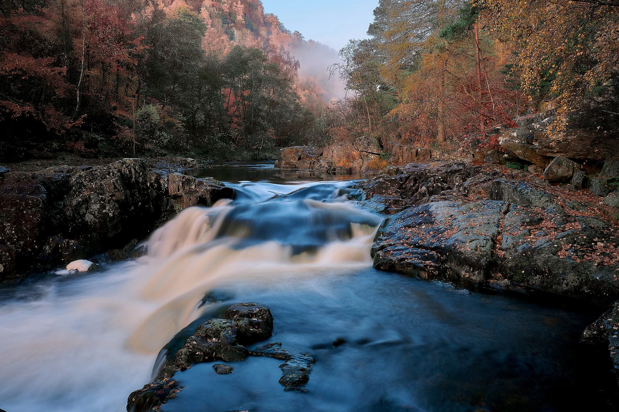 The water slips over the rocks in a soft, silken flow, captured through long exposure to reveal the calm hidden within movement. Around the river, the forest glows with the warm colours of the season — deep reds, golds, and burnt orange — while a gen