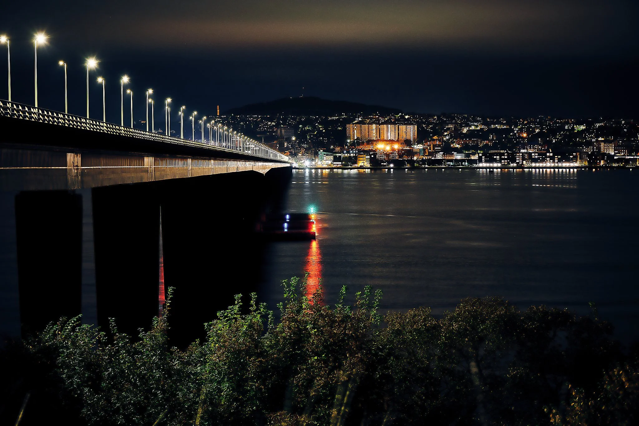 The Tay Road Bridge stretches across the water like a glowing thread, its line of lights guiding the eye toward the bright heart of Dundee. Night settles softly over the river, turning the surface into a dark mirror that reflects the colours of the b