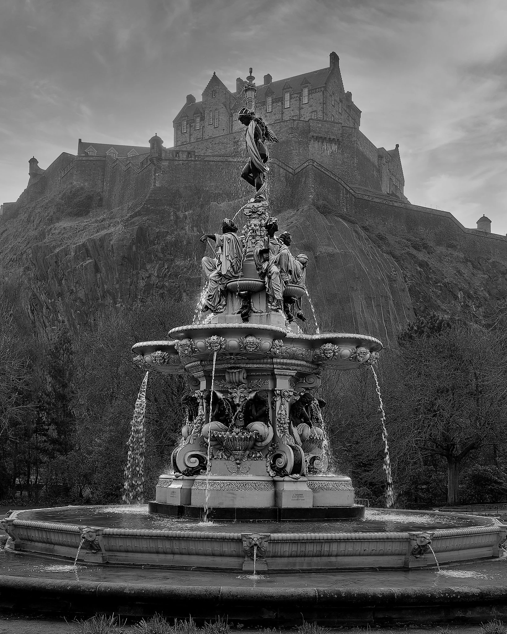 Captured at Ross Fountain beneath the imposing rise of Edinburgh Castle, this black‑and‑white composition distils the city’s grandeur into pure form and contrast. The ornate figures of the fountain stand in quiet dialogue with the fortress above, the