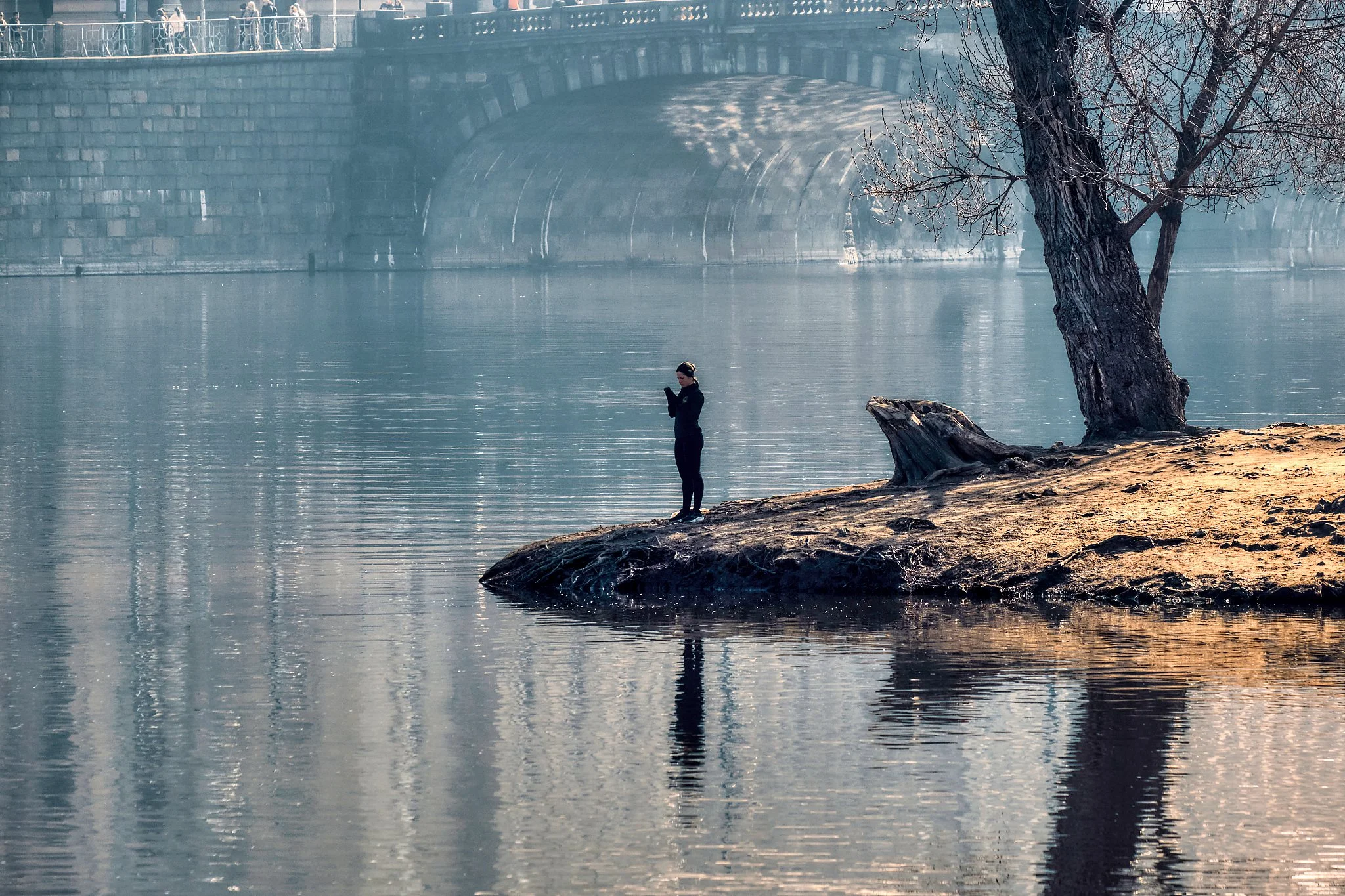 A moment of stillness settles on the banks of the Vltava, where a young woman stands alone at the water’s edge, absorbed in her own quiet world. The river mirrors everything around her—the bare branches of a winter tree, the soft curve of the shoreli