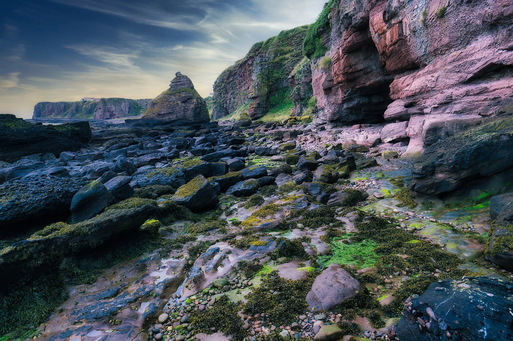 Along the rugged coastline of Auchmithie, the tide has slipped away to reveal a world shaped by time and weather. Wet boulders glisten under a soft, shifting sky, their surfaces brushed with green algae and the delicate sheen of retreating waves. Beh