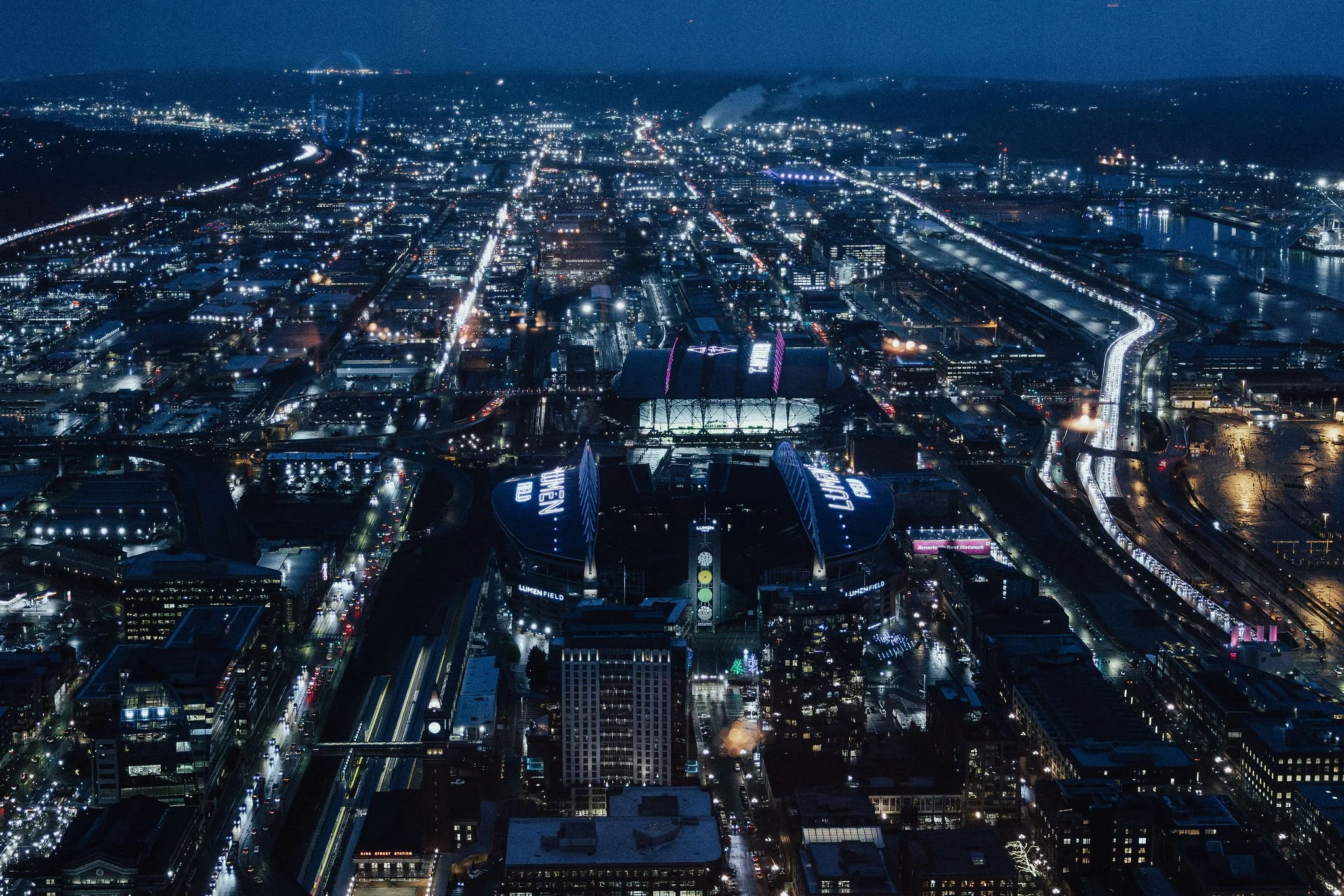 Nighttime aerial view of downtown Seattle featuring Lumen Field, illuminated skyscrapers, city streets, and urban buildings with the backdrop of the dark sky and distant horizon.