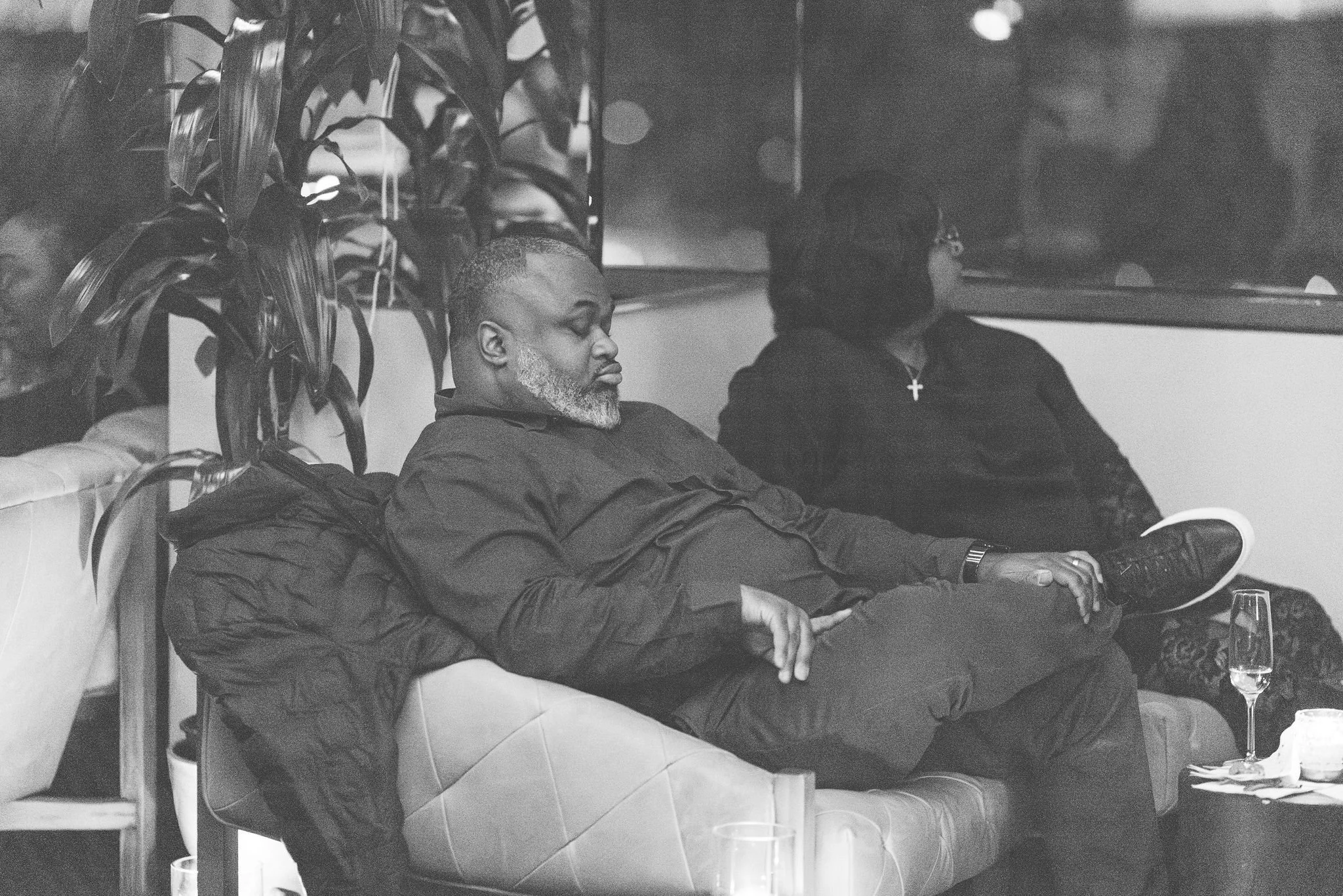 A black and white photo of two people sitting on a couch in a dimly lit room. One person with a beard and short hair appears to be looking at a cell phone, while the other person is facing away and is wearing a cross necklace.