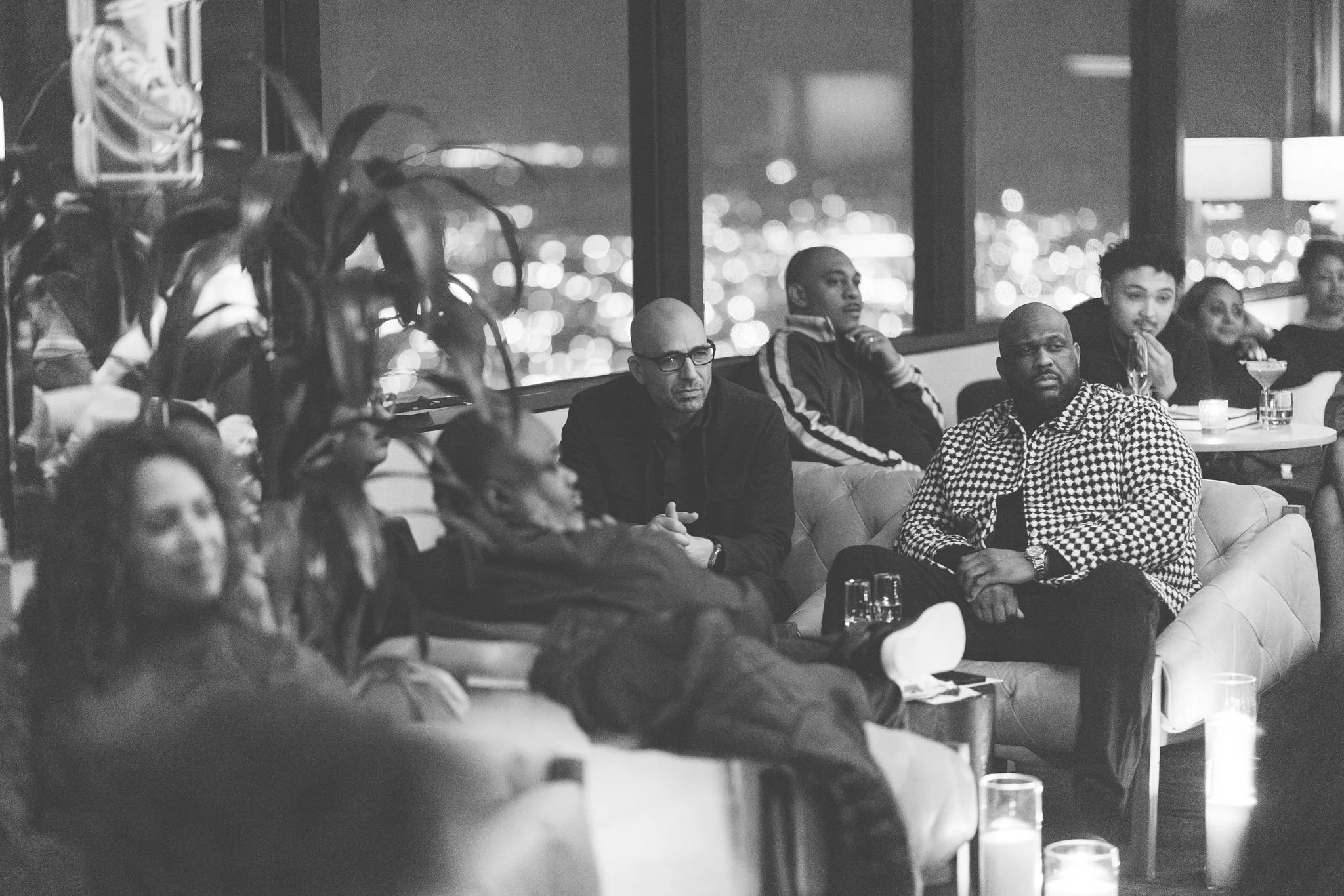 A group of diverse people sitting on sofas and chairs in a modern lounge or bar, watching something intently, with city lights visible through large windows at night.