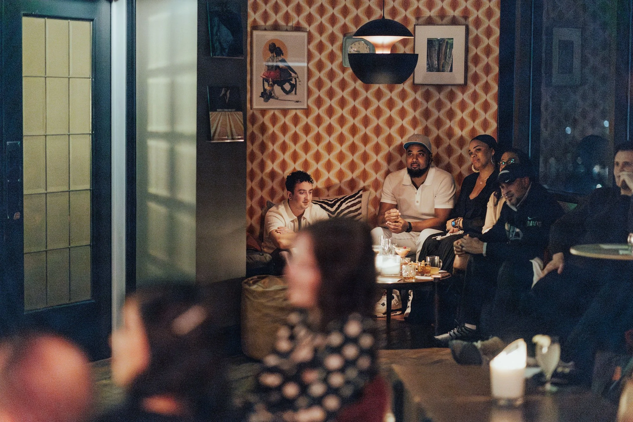 A group of people sitting on a couch with drinks in a dimly lit room with vintage decor and patterned wallpaper, during a social gathering.