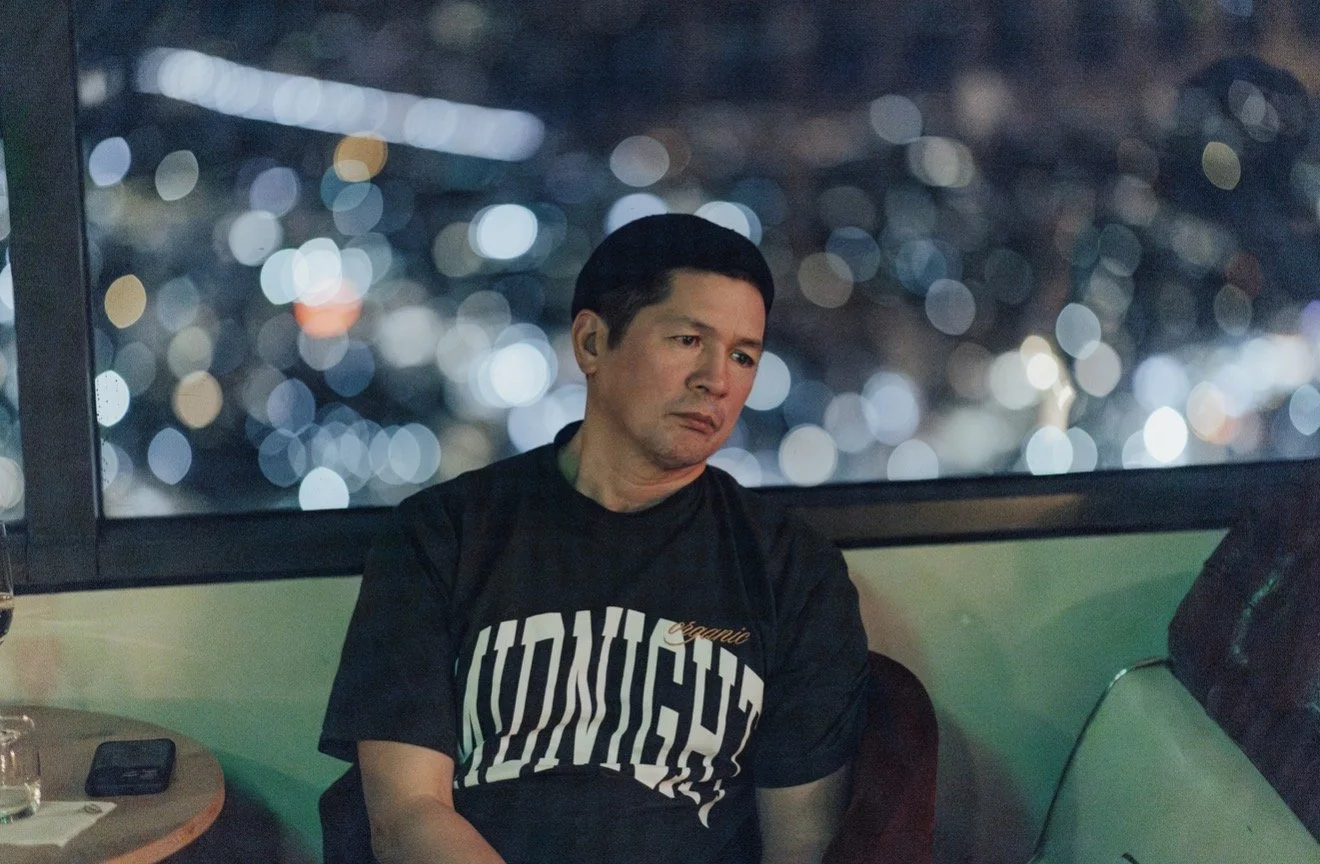A man sitting at a table in a restaurant or bar, looking out the window with a cityscape glowing with lights in the background at night.
