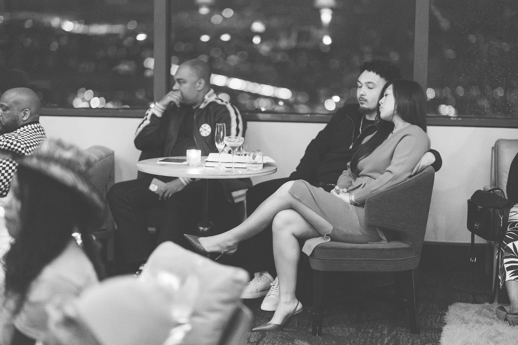 Black and white photo of three people sitting at a table and chair in a bar or lounge. The woman in the center is sitting with her legs crossed, dressed in a professional outfit, and has her head leaning on the man's shoulder next to her. The man is 