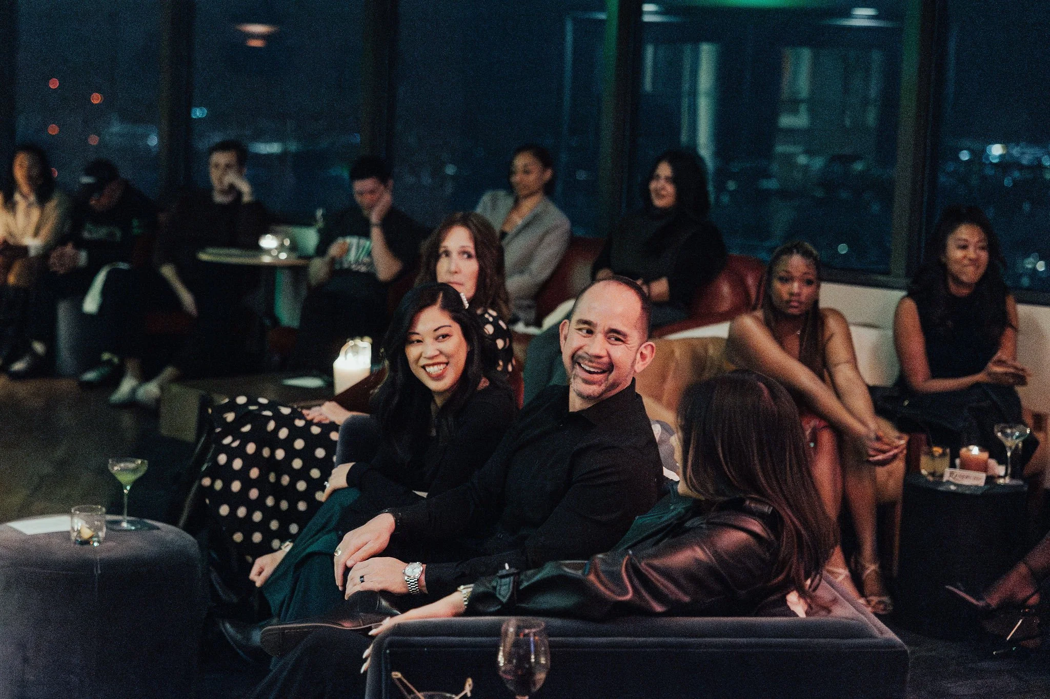 People sitting in a dimly lit lounge or bar, some smiling and looking at the camera, with drinks on tables and large windows in the background showing a city at night.
