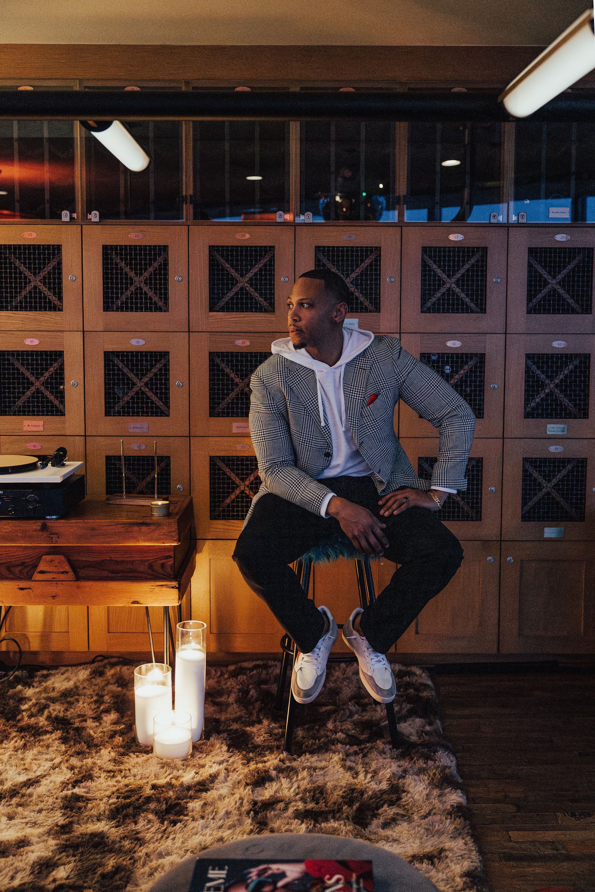 A man sitting on a stool in a room with wooden cabinets behind him, a turntable on a side table, and candles on the floor in front of him.