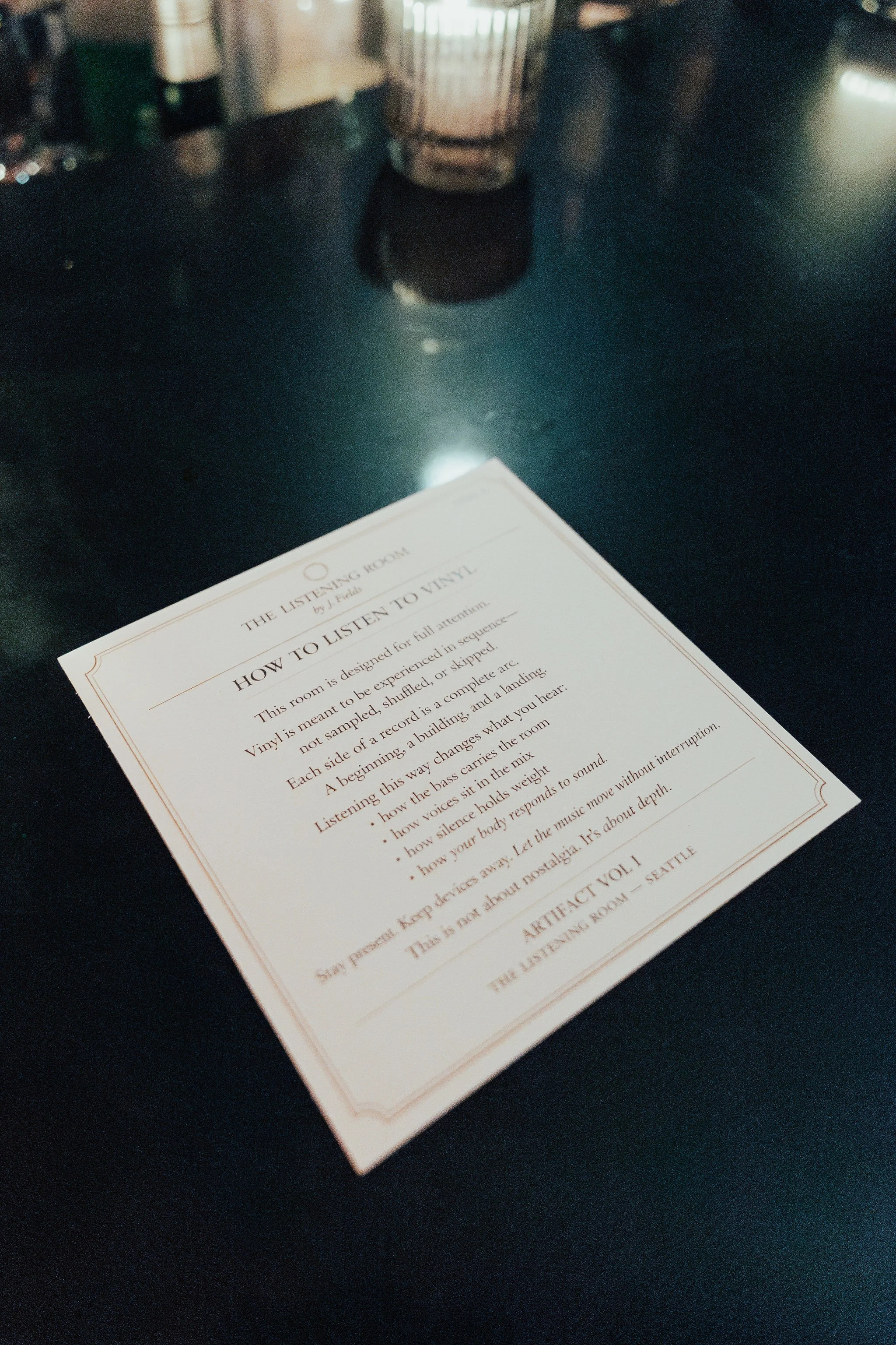A printed sheet of paper titled 'The Listening Room' with instructions for listening to vinyl records, placed on a dark table surface. There is a blurred background with bottles and a decorative item.