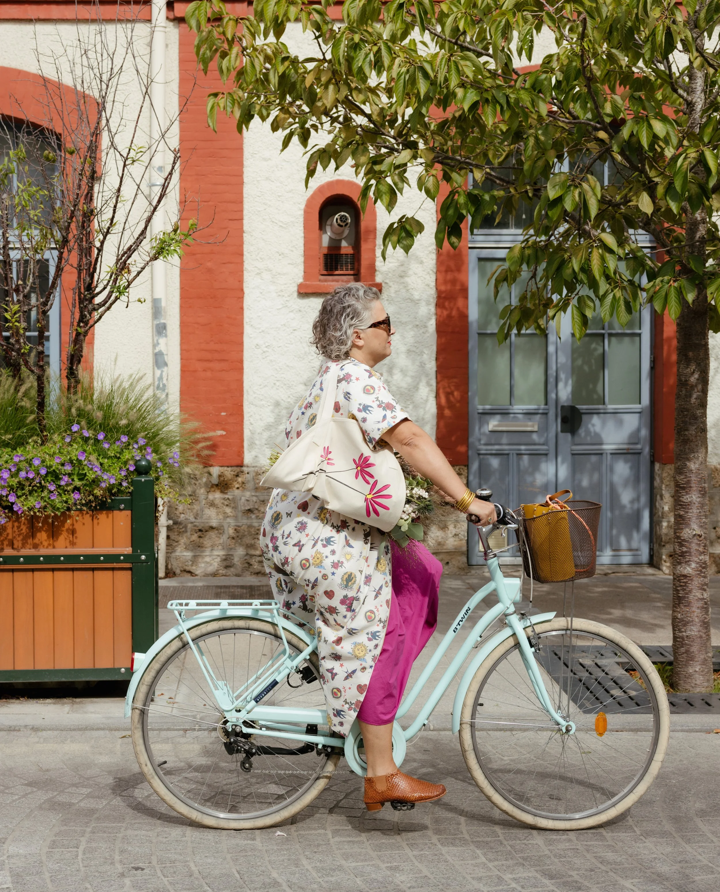 Une femme âgée porte un chemisier blanc à motifs floraux, des pantalons roses, des lunettes de soleil, et des bracelets dorés, elle pédale à vélo avec un panier en osier à l'avant, dans une rue bordée d'arbres et de bâtiments colorés.