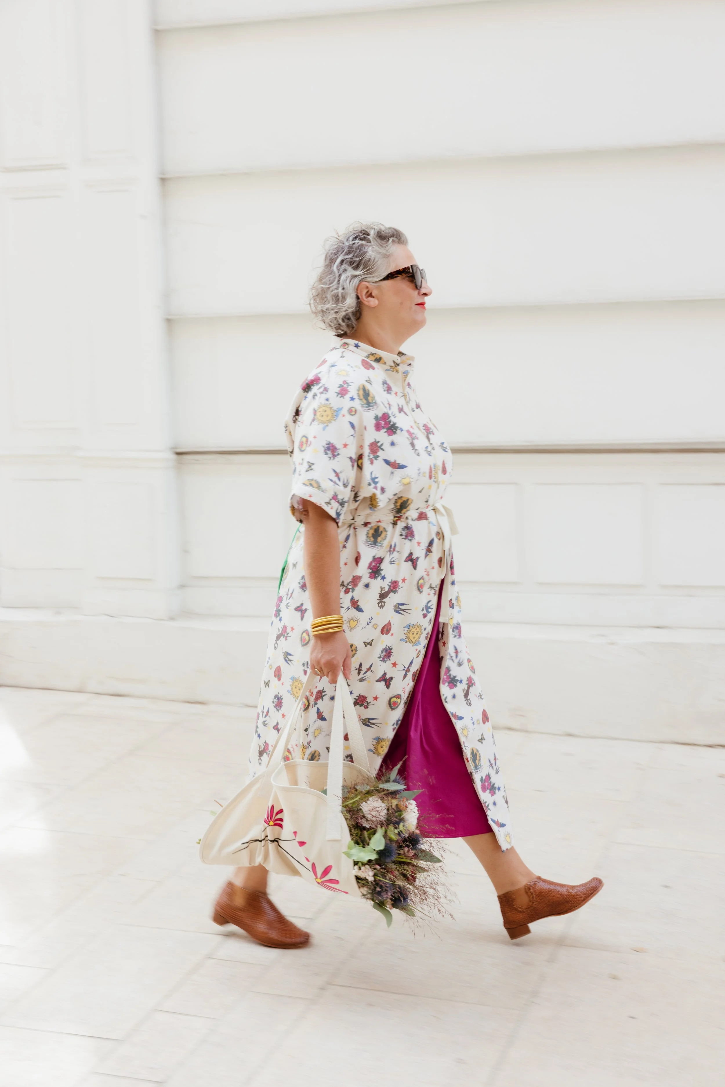 Femme portant une robe longue à motifs floraux, portant des lunettes de soleil, et tenant des sacs, marchant à l'extérieur près d'un mur blanc.