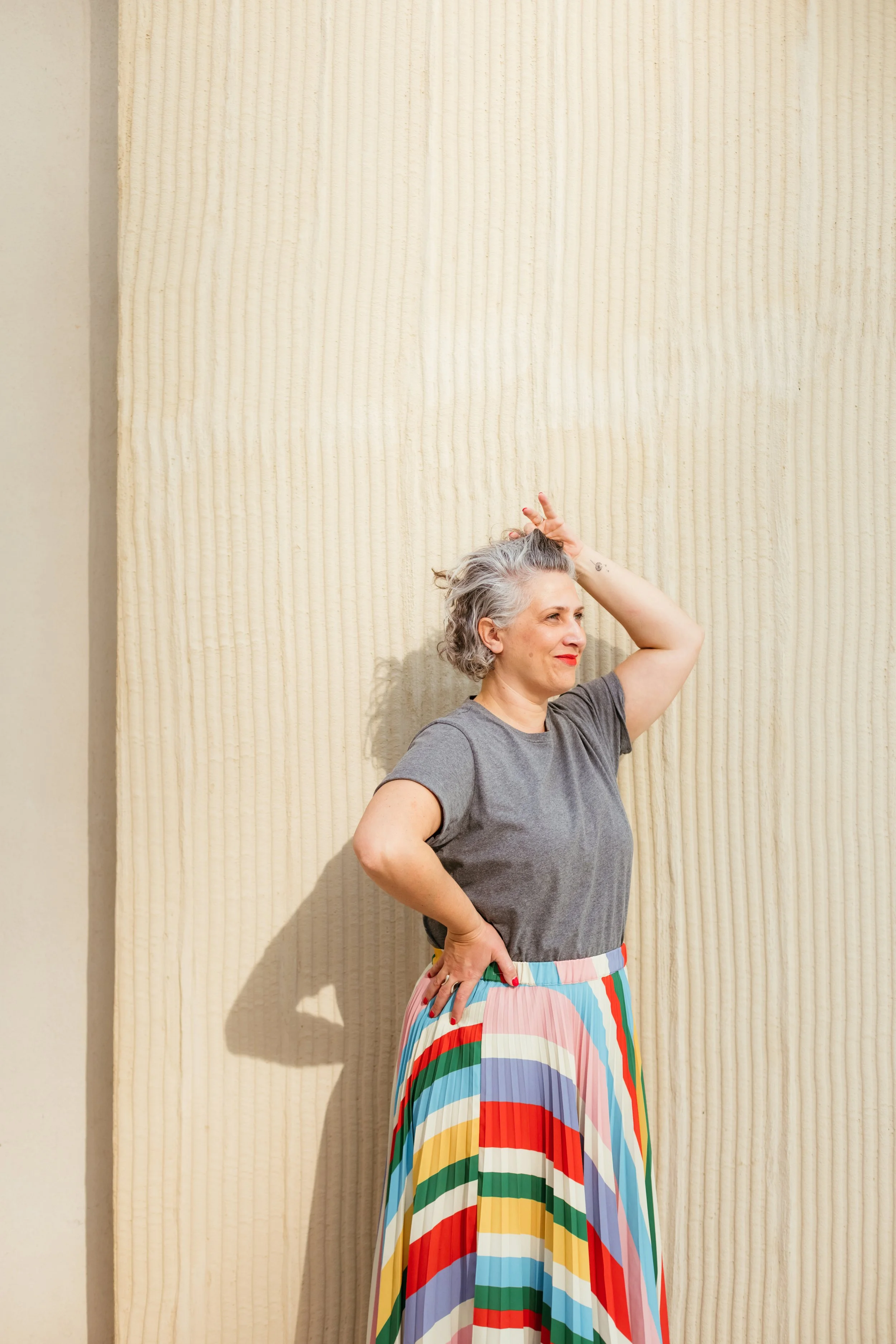 Une femme avec des cheveux gris, portant un t-shirt gris et une jupe longue à motifs arc-en-ciel, se tient devant un mur en bois clair, levant la main droite pour toucher ses cheveux.
