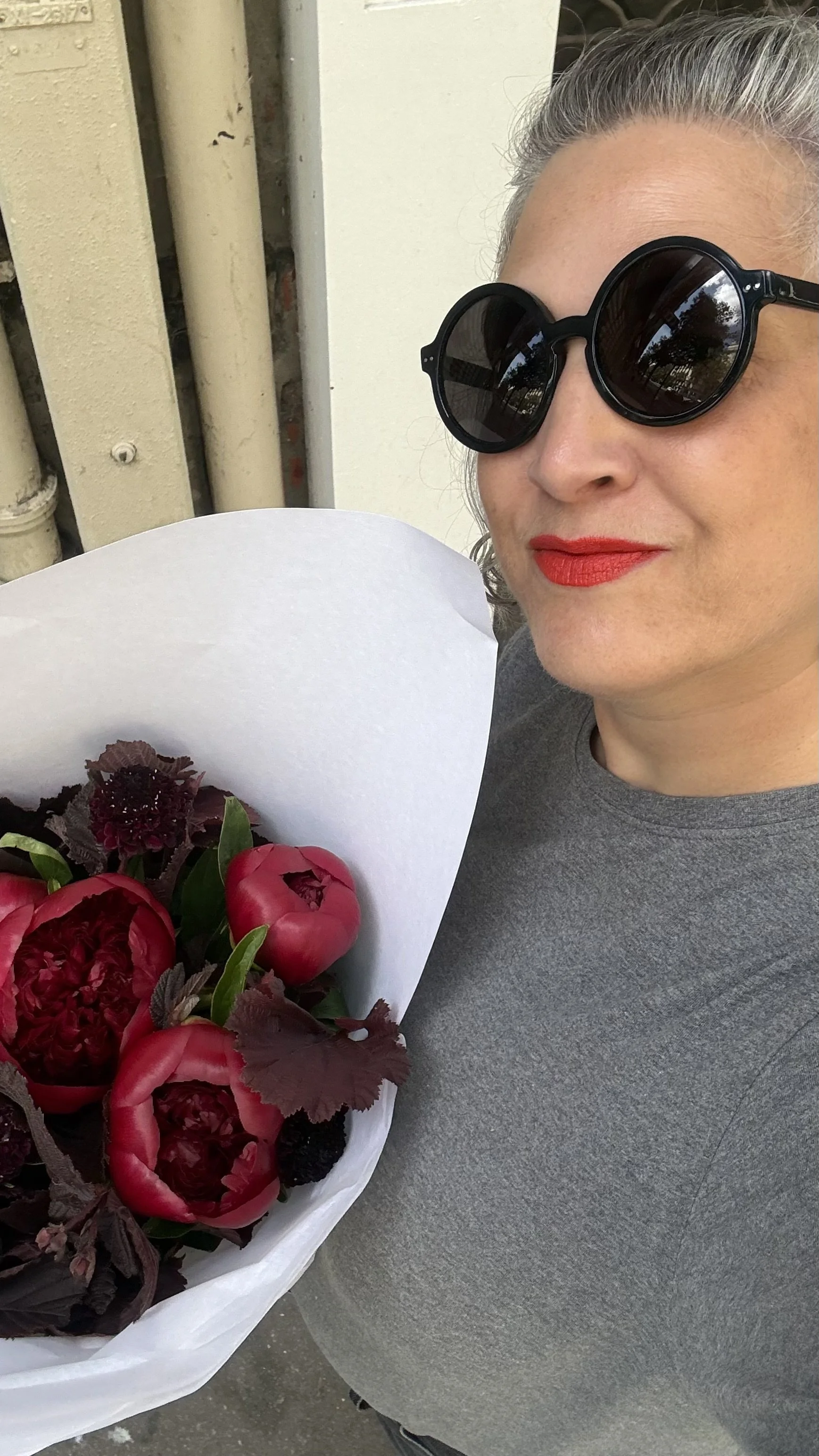 Une femme portant des lunettes de soleil noires, souriante, tient un bouquet de fleurs rouges et roses enveloppé dans du papier blanc.