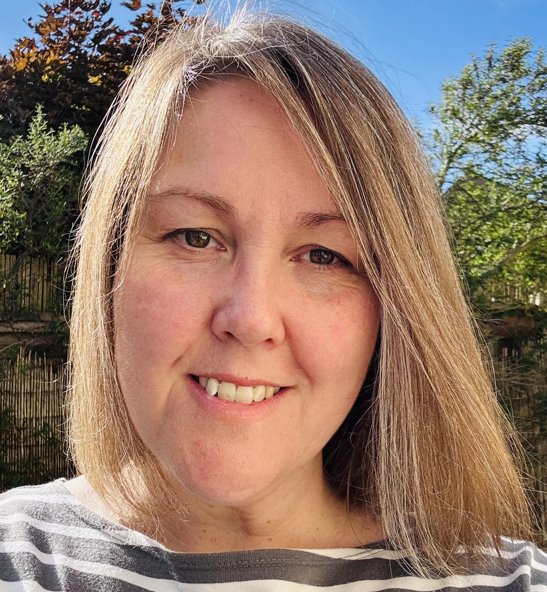 Close-up of a smiling woman with light brown hair outdoors, wearing a striped shirt, with greenery and blue sky in the background.