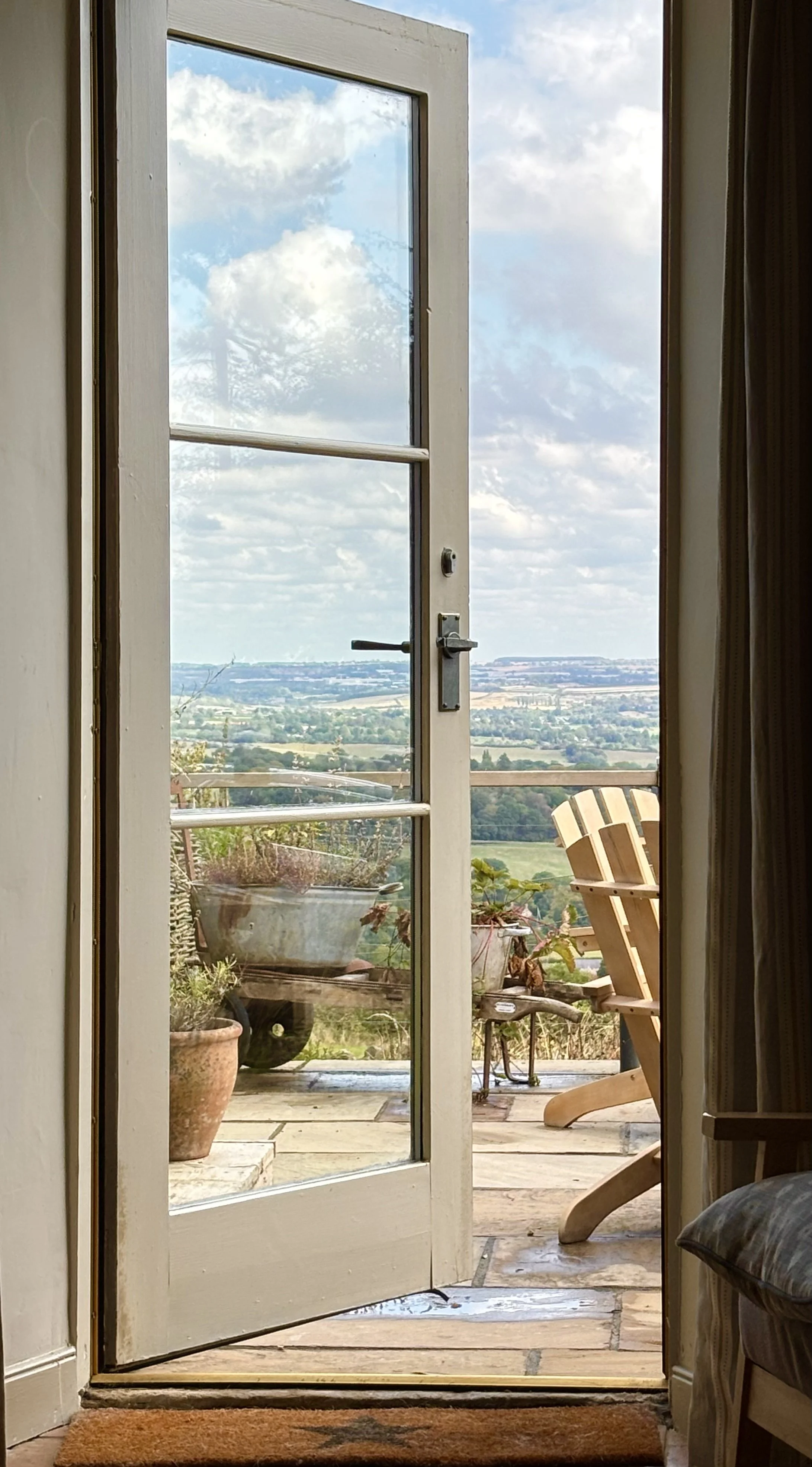A view through a glass-paneled door opening to a balcony with potted plants, a wooden chair, and a scenic landscape of fields and trees under a partly cloudy sky.
