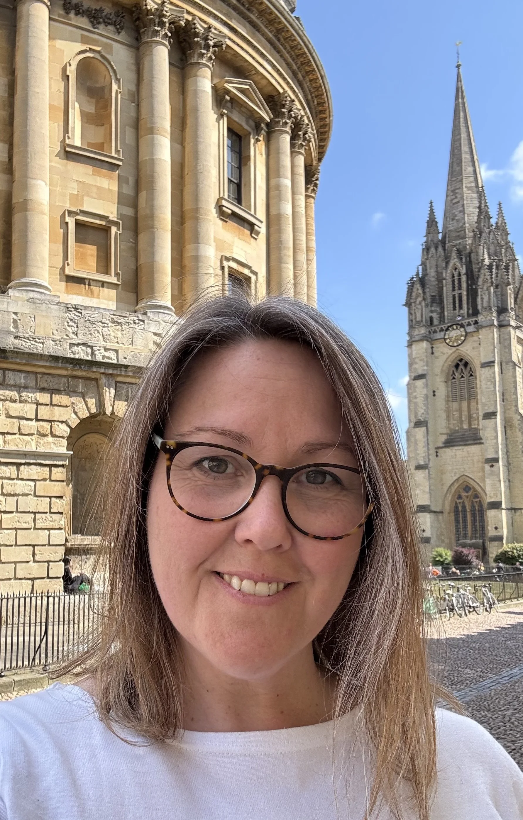 A woman wearing glasses, smiling, taking a selfie in front of a historic church with large stone columns, gothic windows, and a tall spire, on a clear day.