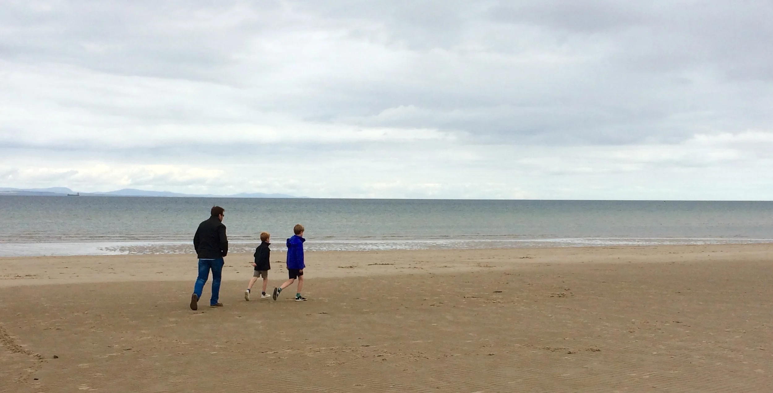 A man and two children walking on a sandy beach near the water under a cloudy sky.