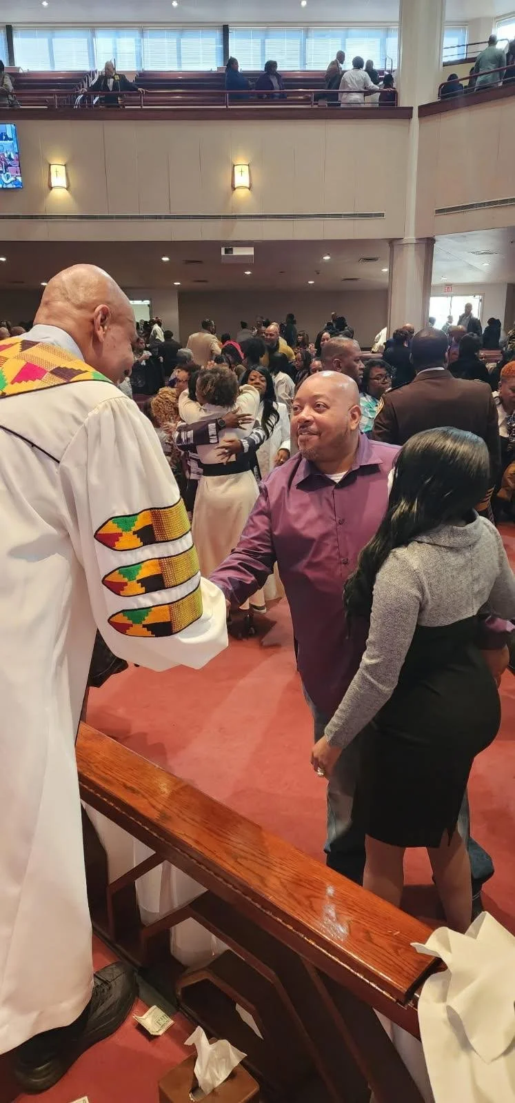 A group of people in a church or community hall, with some hugging, shaking hands, and engaging in conversations during a gathering or service. The room is filled with attendees, some seated upstairs and others standing or congregating on the main fl