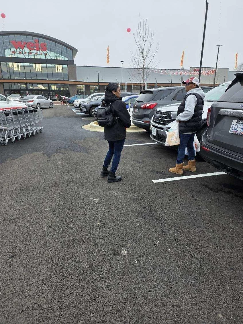 Two women talking in a parking lot outside a Wegmans supermarket, with shopping carts and parked cars around.
