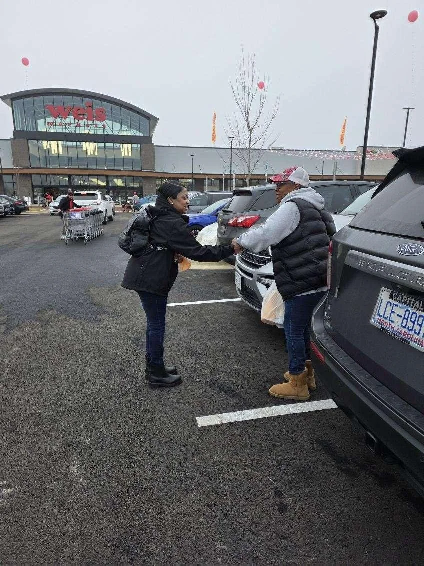 A woman and a man shaking hands in a parking lot outside a Wegmans supermarket on a cloudy day. The woman is wearing a black jacket and boots, and the man is dressed in a gray hoodie, black puffy vest, jeans, and tan boots. There are cars parked arou