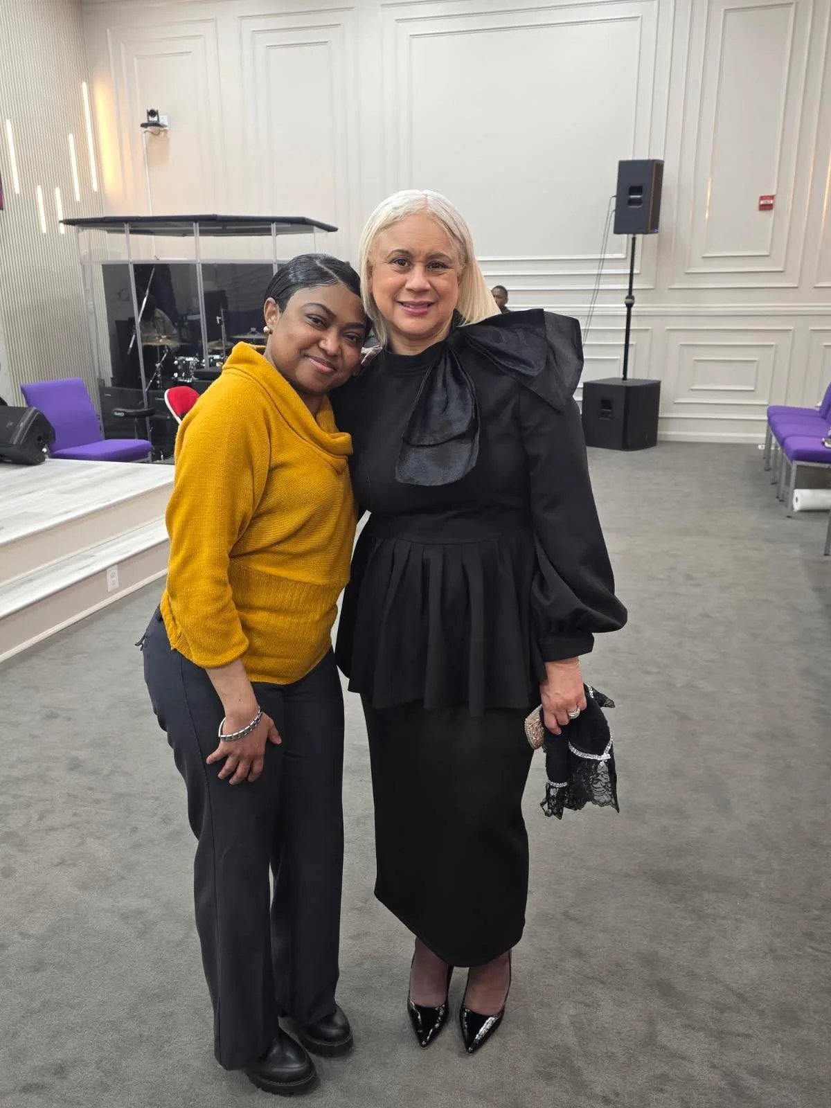Two women standing side by side in a room with purple chairs and musical instruments in the background.