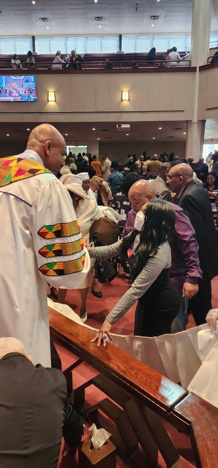 People gathering in a church during a religious service or event, engaging in prayer or worship, with some dressed in ceremonial robes and others in casual or formal clothing, inside a spacious church with a balcony and a television screen.