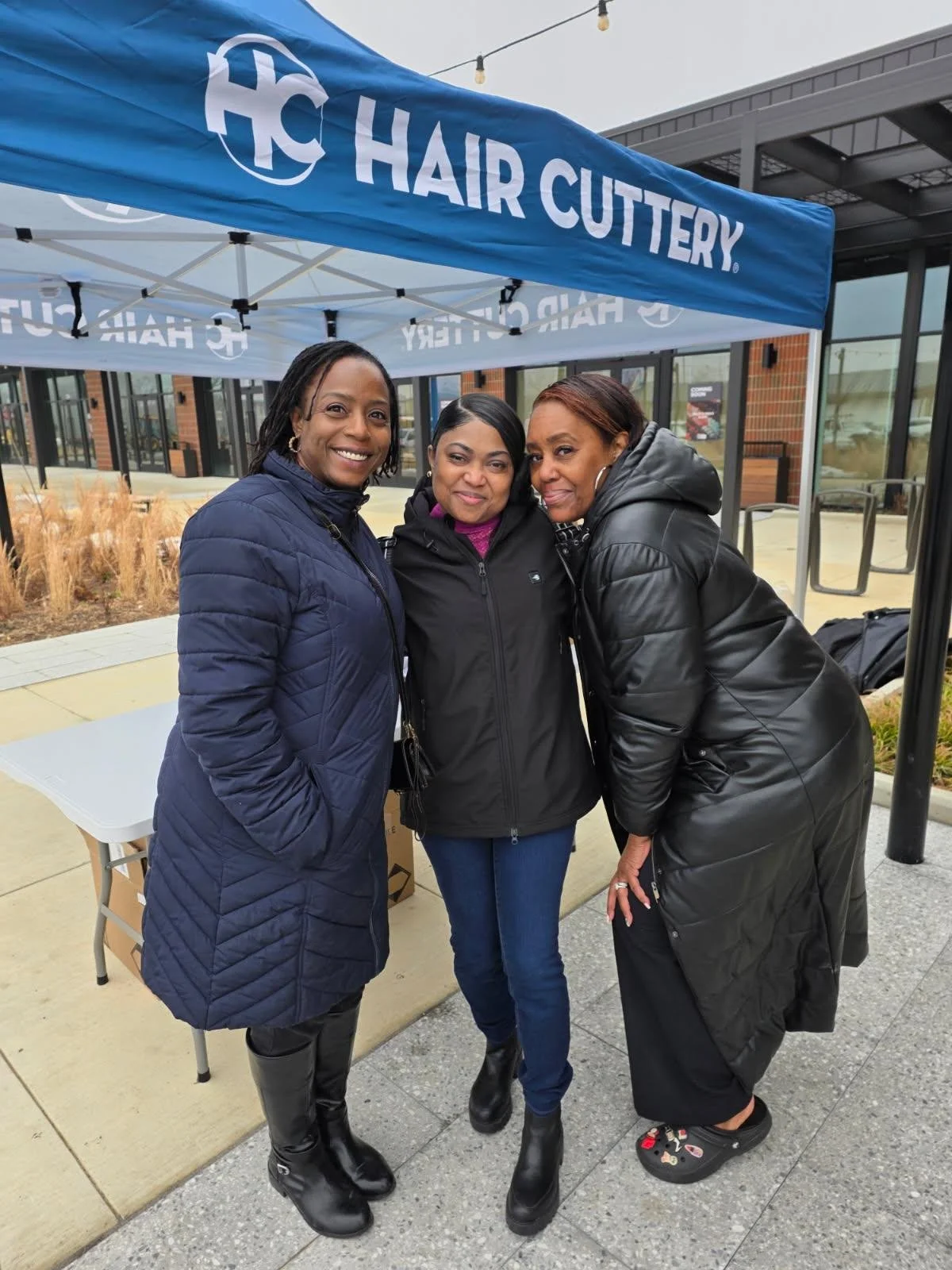 Three women standing together, smiling, under a blue canopy with white text that reads 'HAIR CUTTERY.' They are outside a commercial building, dressed in dark winter jackets and boots, and appear to be happy.